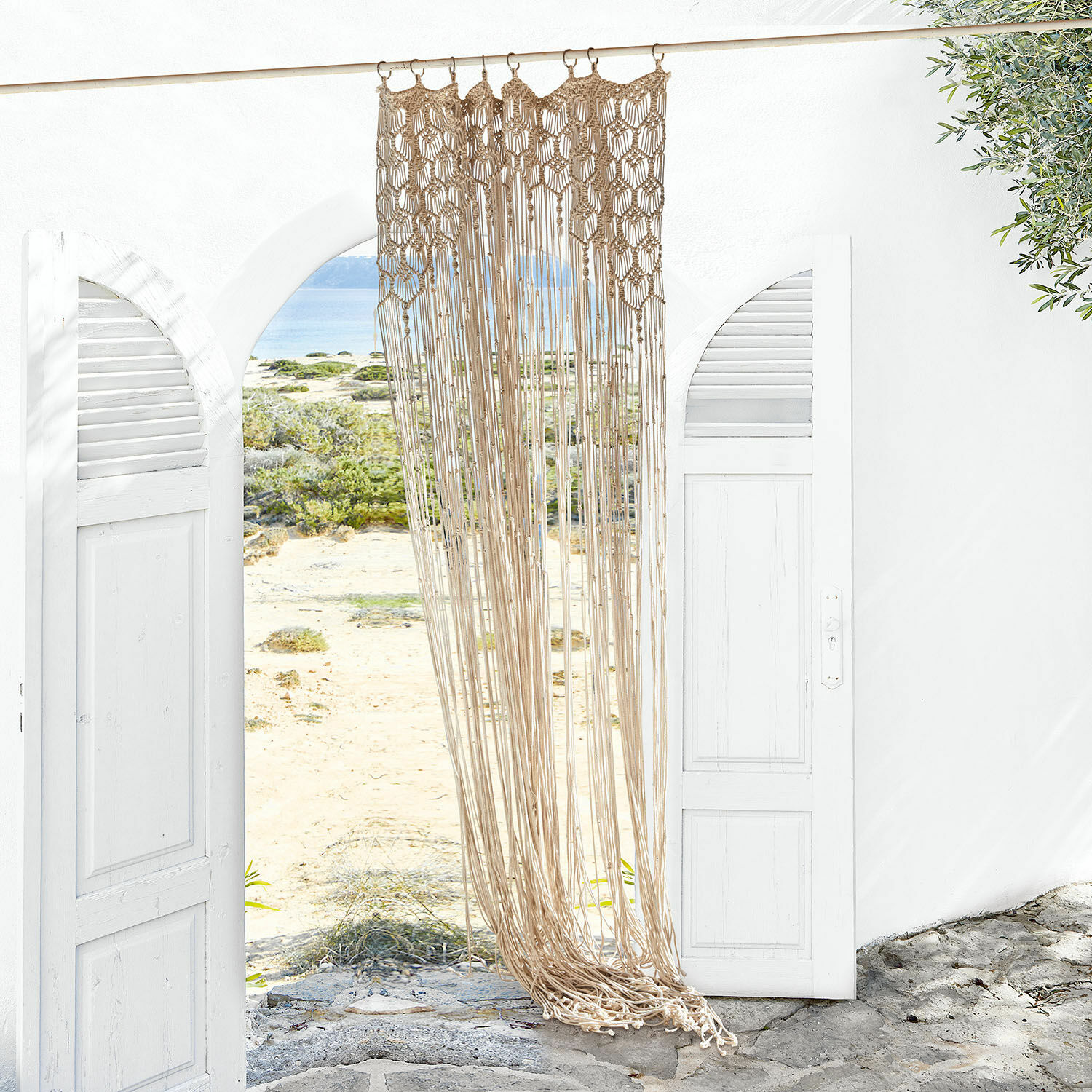 Cortina Manville Macramé beige de algodón colgado en una entrada blanca abierta con vista a la playa y el mar
