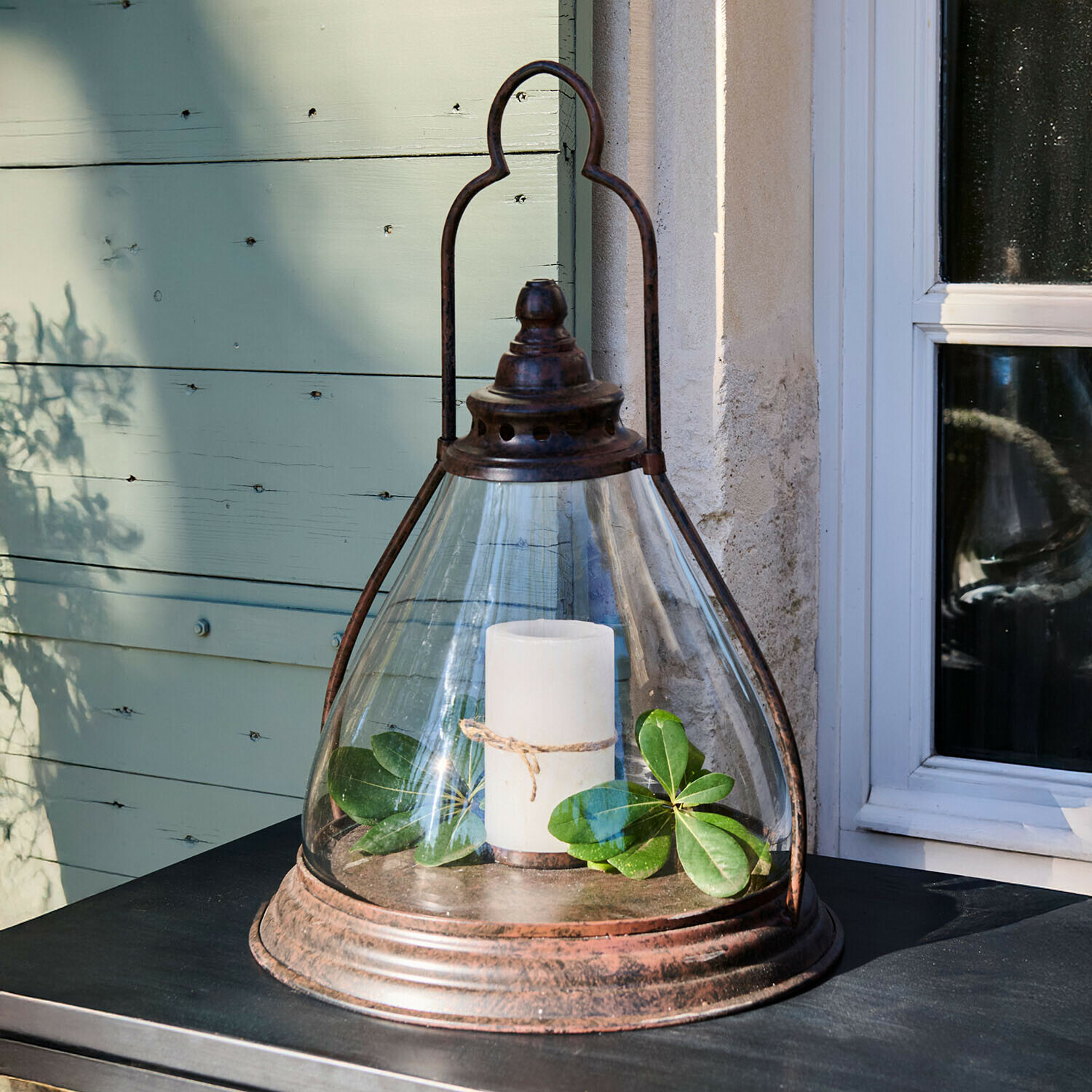 Linterna de vidrio con marco metálico oxidado, vela blanca y hojas verdes sobre mesa exterior