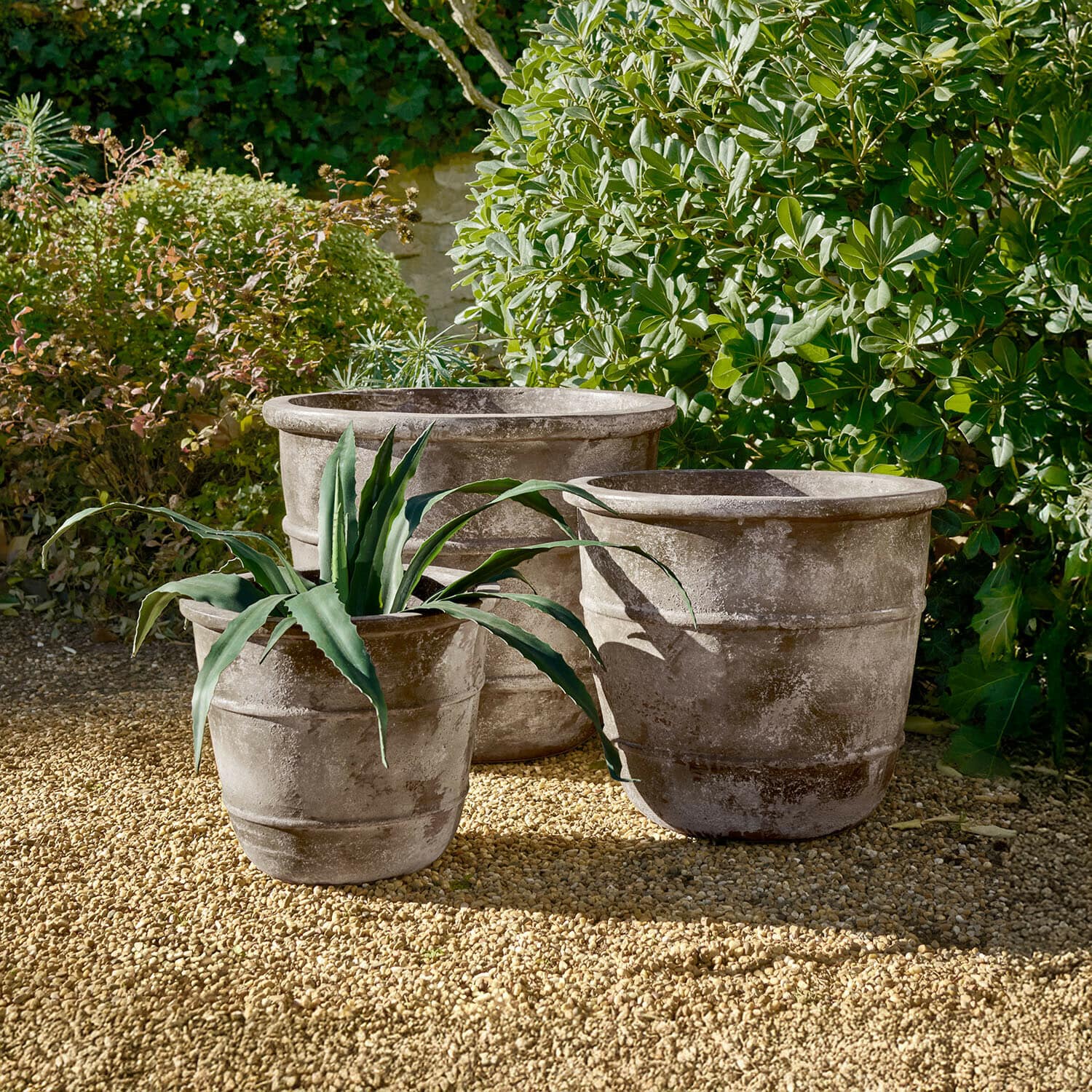 Set de 3 maceteros Arlivonne Tres grandes macetas de piedra gris sobre grava en jardín con plantas verdes alrededor
