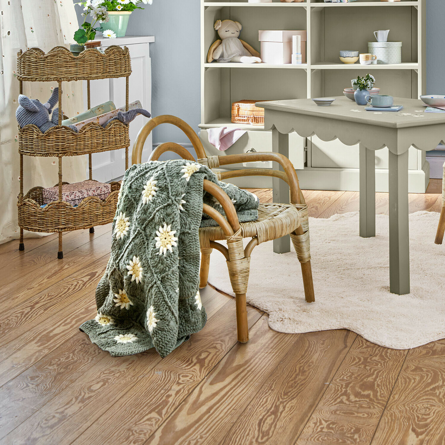 Silla infantil Tinytreasure Manta gris tejida con flores blancas sobre silla de mimbre en habitación infantil luminosa con suelo de madera