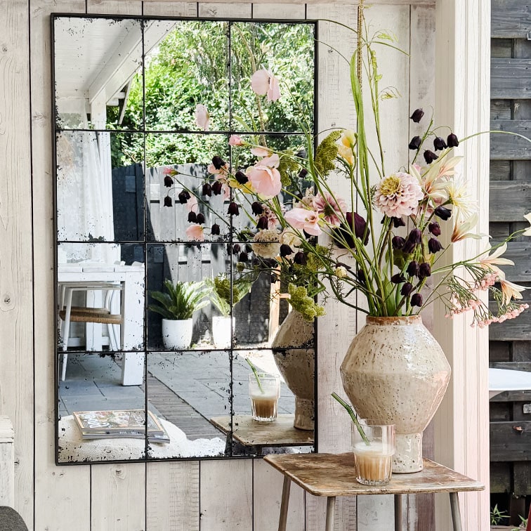 Espejo de pared con paneles que refleja flores en jarrón de cerámica y café sobre mesa de madera rústica