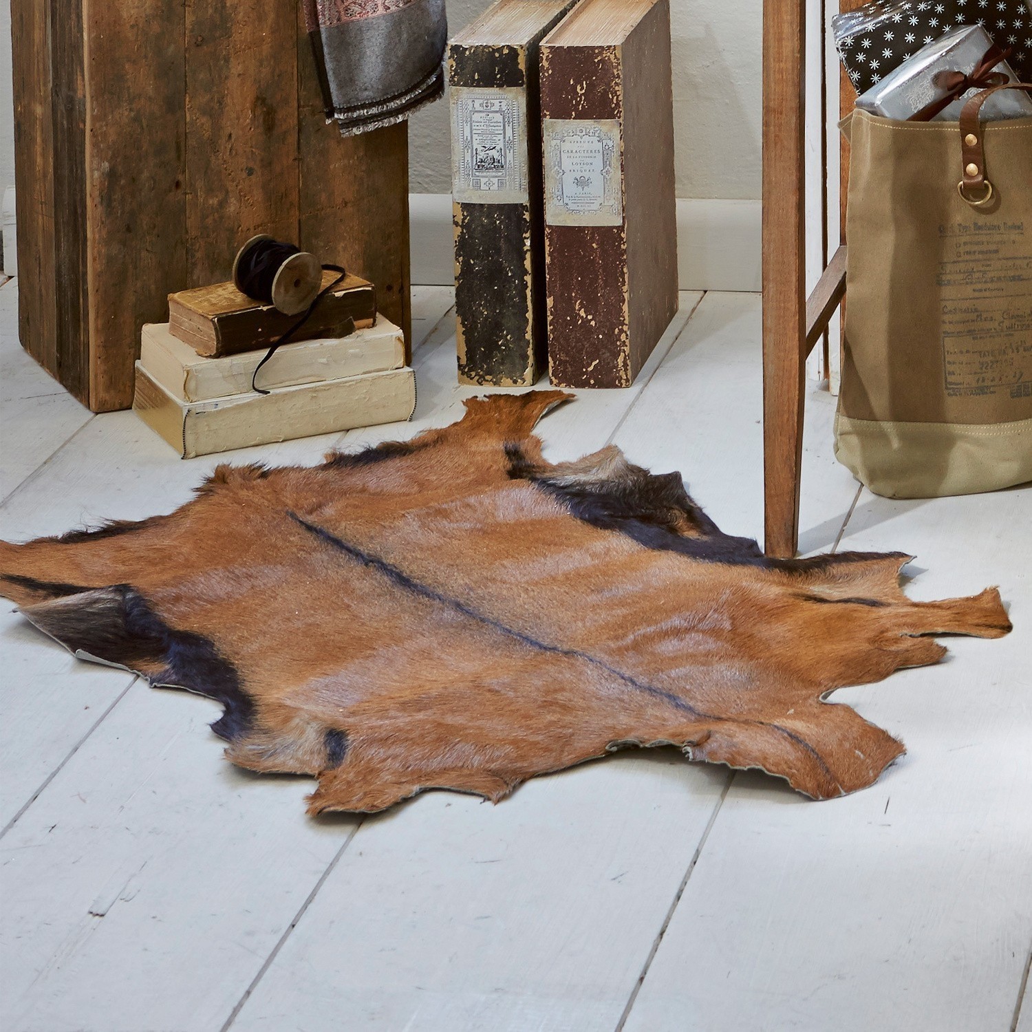 Alfombra de piel de cabra Piedro Piel natural marrón sobre suelo de madera blanca con libros antiguos y muebles rústicos