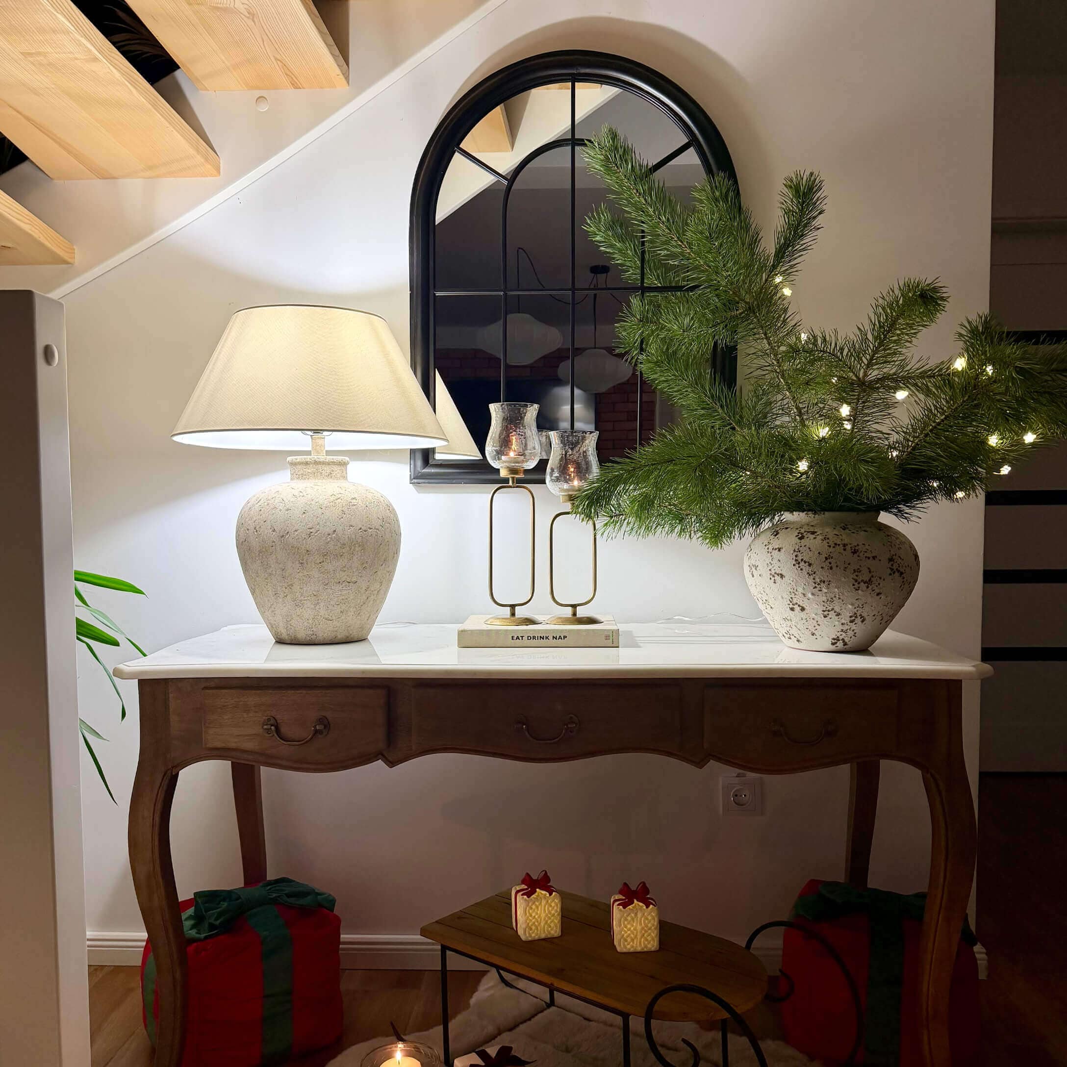 Mesa de madera con superficie blanca, lámpara de cerámica, espejo y decoración navideña con ramas verdes y luces