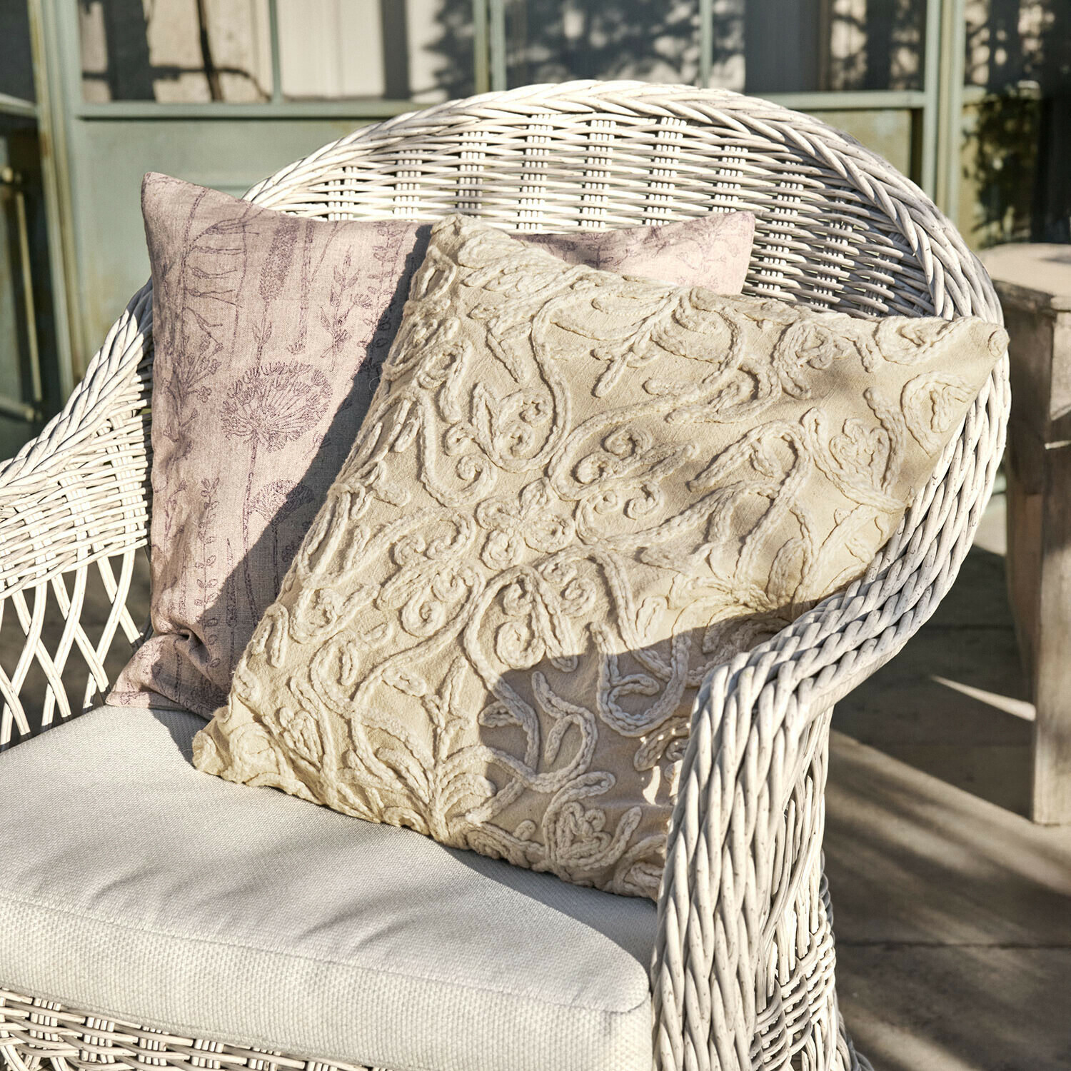Set de 2 fundas de cojín Floraine Dos cojines decorativos beige y rosa sobre un sillón de mimbre blanco en una terraza soleada