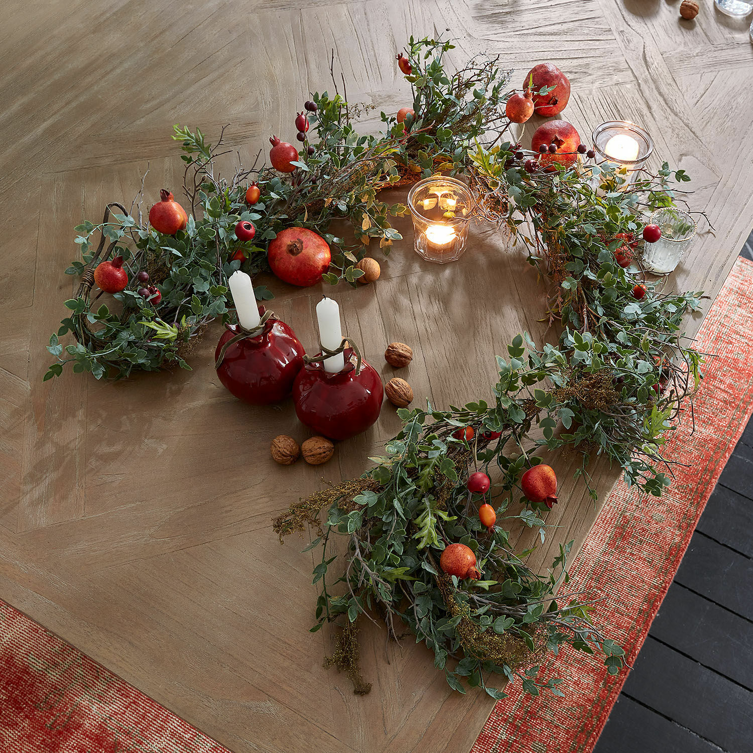 Guirnalda Karidah Corona decorativa con hojas verdes y frutos rojos sobre mesa de madera con velas encendidas