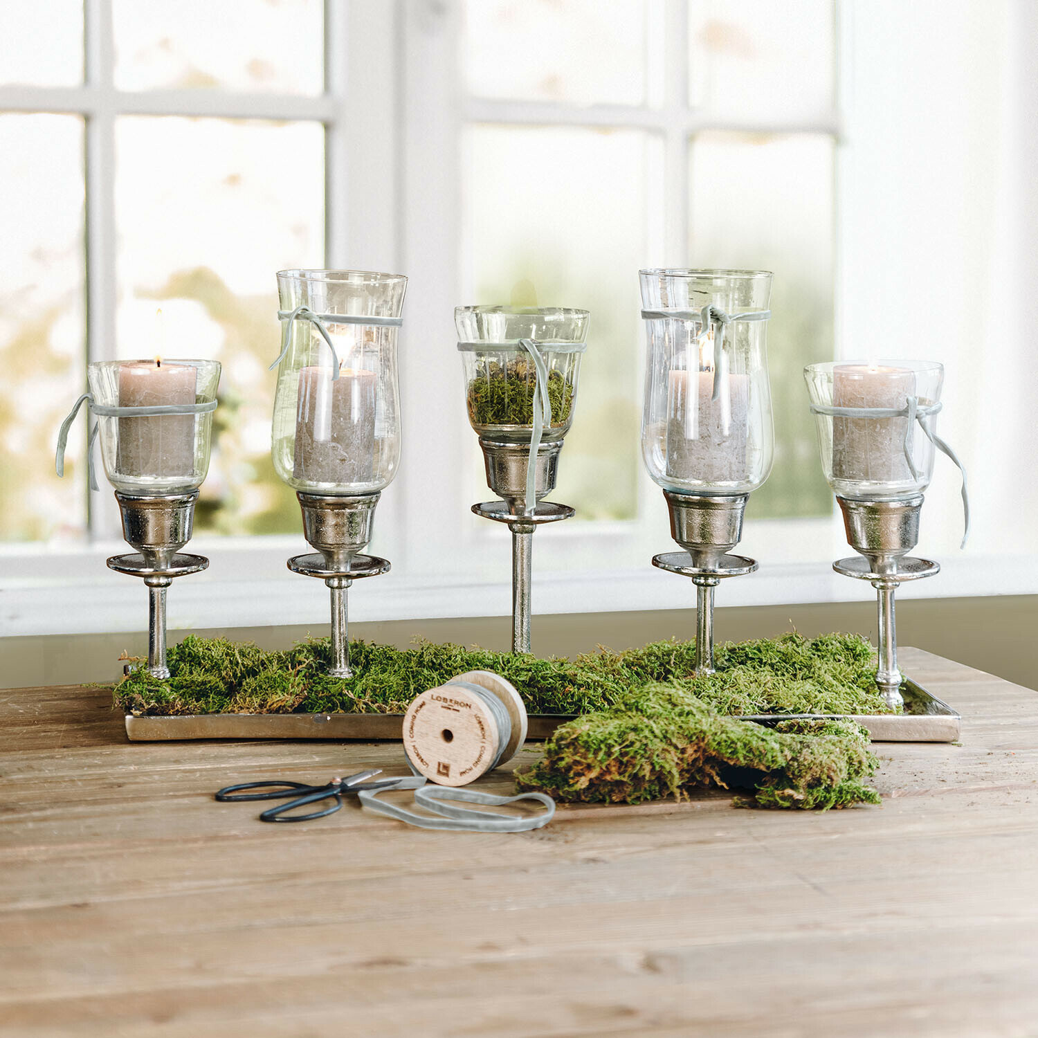 Conjunto de cinco portavelas de vidrio con base metálica y musgo verde sobre mesa de madera