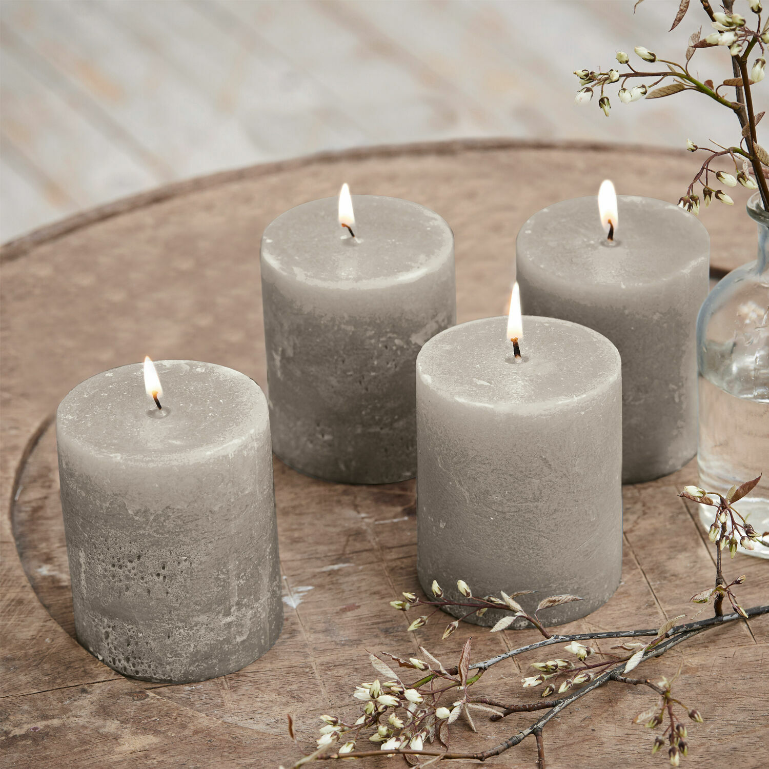 Set de 4 velas Virrans Conjunto de cuatro velas de cera grises sobre mesa de madera con decoración floral, decoración interior