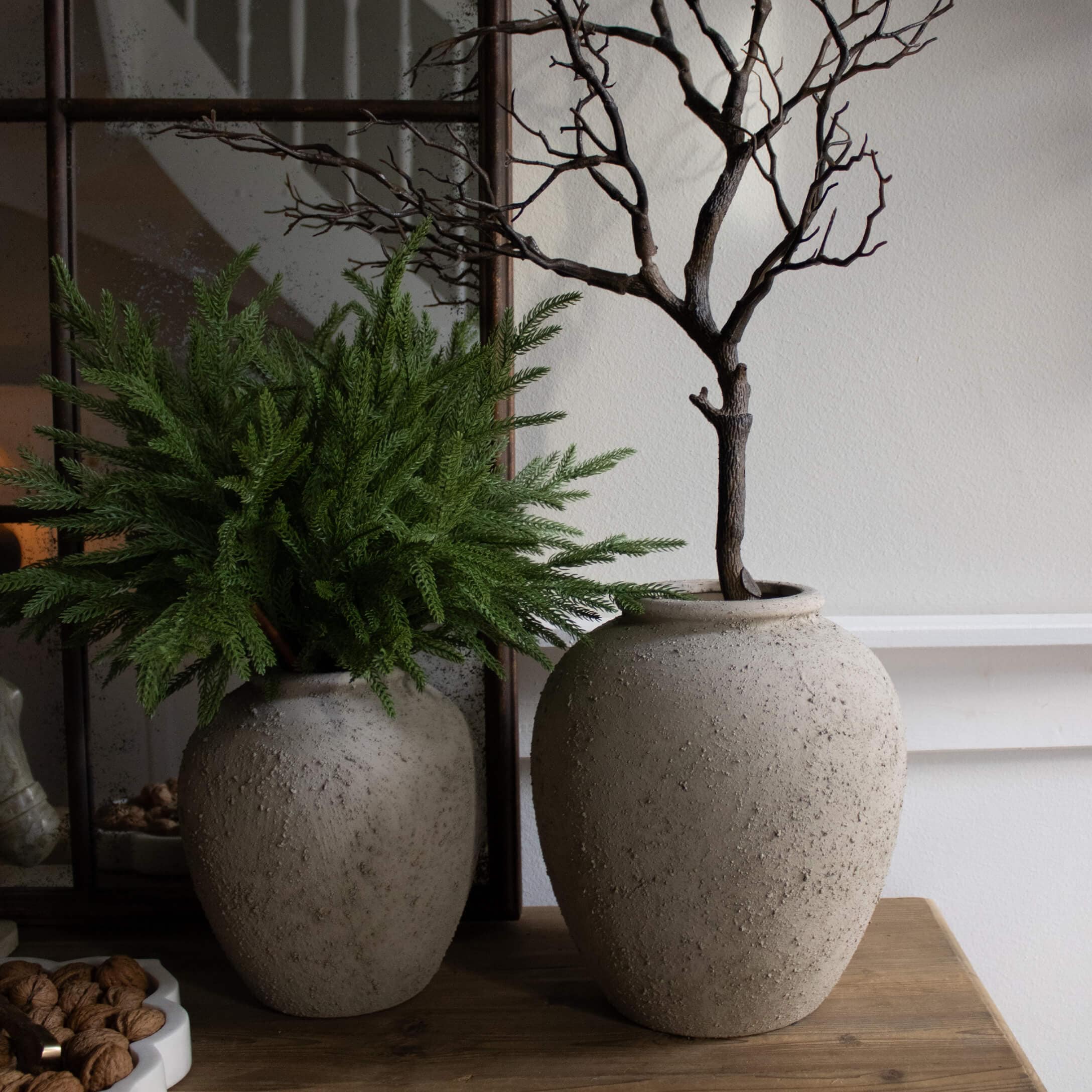 Dos jarrones decorativos de piedra gris sobre mesa de madera con ramas verdes y secas