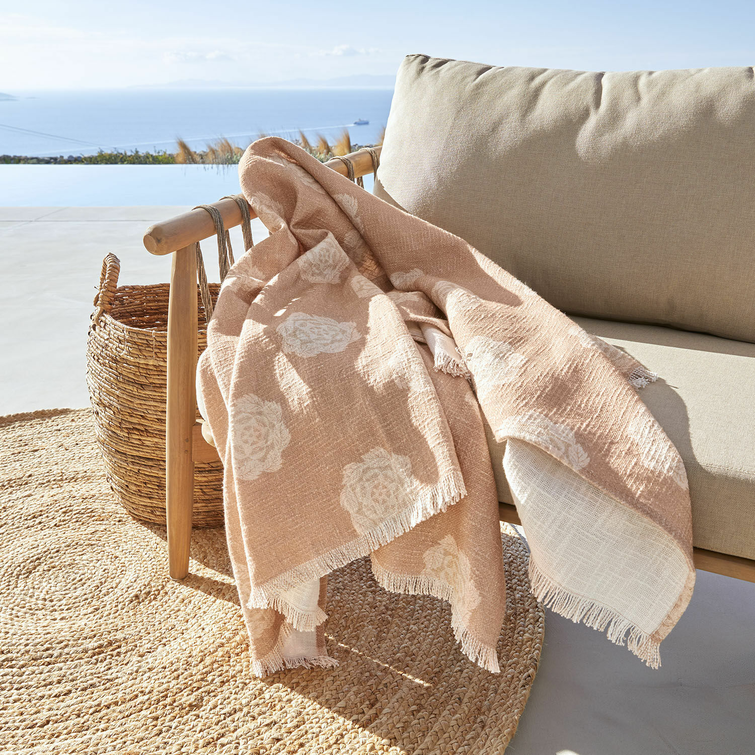 Plaid Belvienne Manta beige con diseño floral blanco de tela suave sobre sillón de madera en terraza con vista al mar