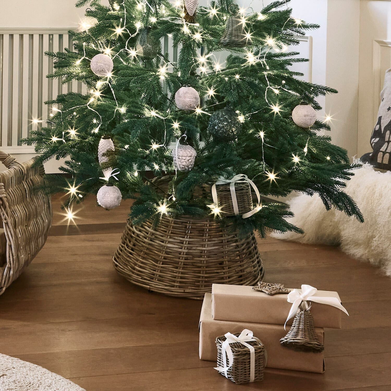 Cesta para árbol de Navidad Kiente Árbol de Navidad en base de mimbre con adornos naturales y regalos envueltos en papel marrón sobre suelo de madera