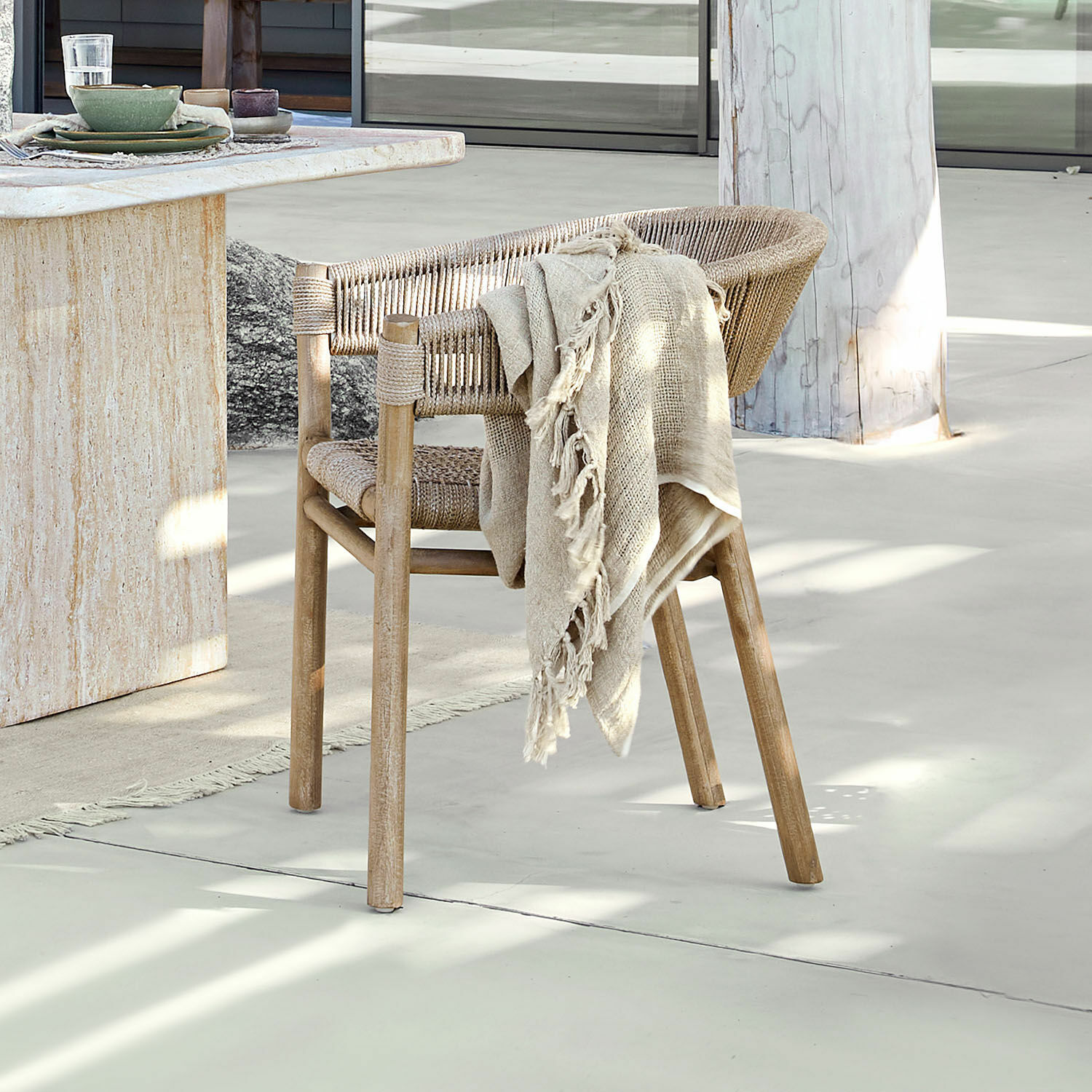 Silla FSC®-100% Garmontique Silla de madera y cuerda natural en color claro con manta beige sobre suelo claro junto a mesa de piedra