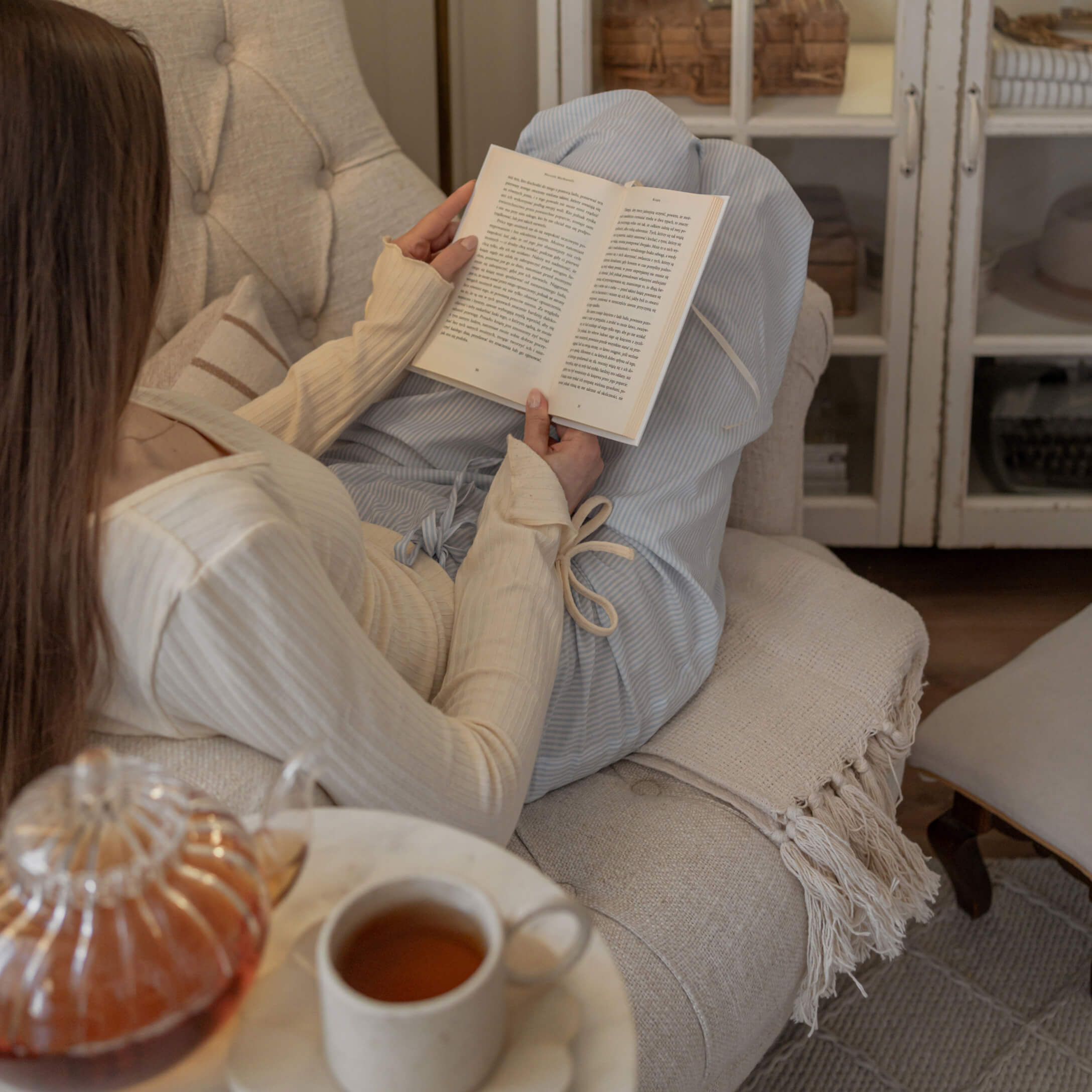 Mujer con suéter crema y pantalones azules leyendo un libro en sofá beige con taza de té en mesa