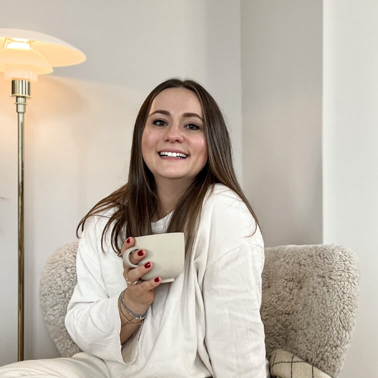 Mujer sonriente con suéter blanco sosteniendo taza beige sentada en sillón suave en interior luminoso