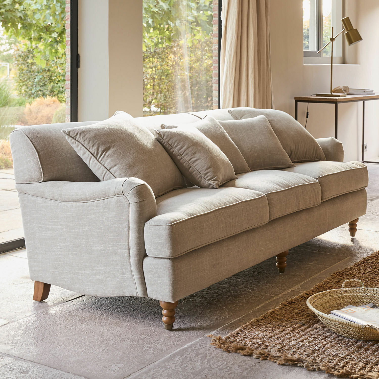 Sofá Isa Sofá beige tapizado con cojines y patas de madera en sala luminosa con alfombra