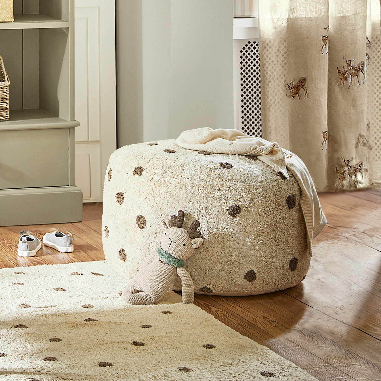 Puf Thivalie Puf beige de material suave con puntos sobre suelo de madera, juguete de peluche y alfombra