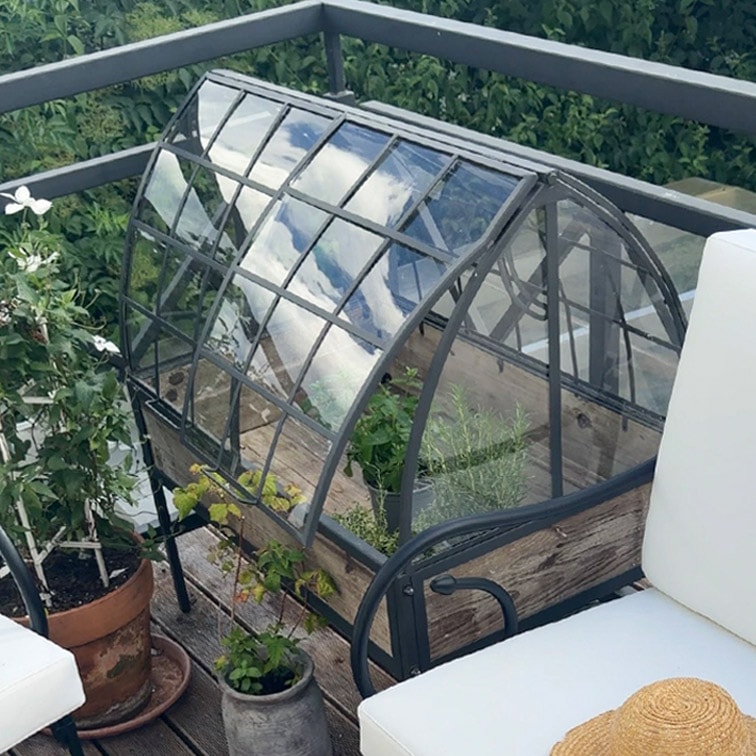 Pequeño invernadero de jardín con estructura metálica y base de madera en terraza con plantas y muebles de exterior