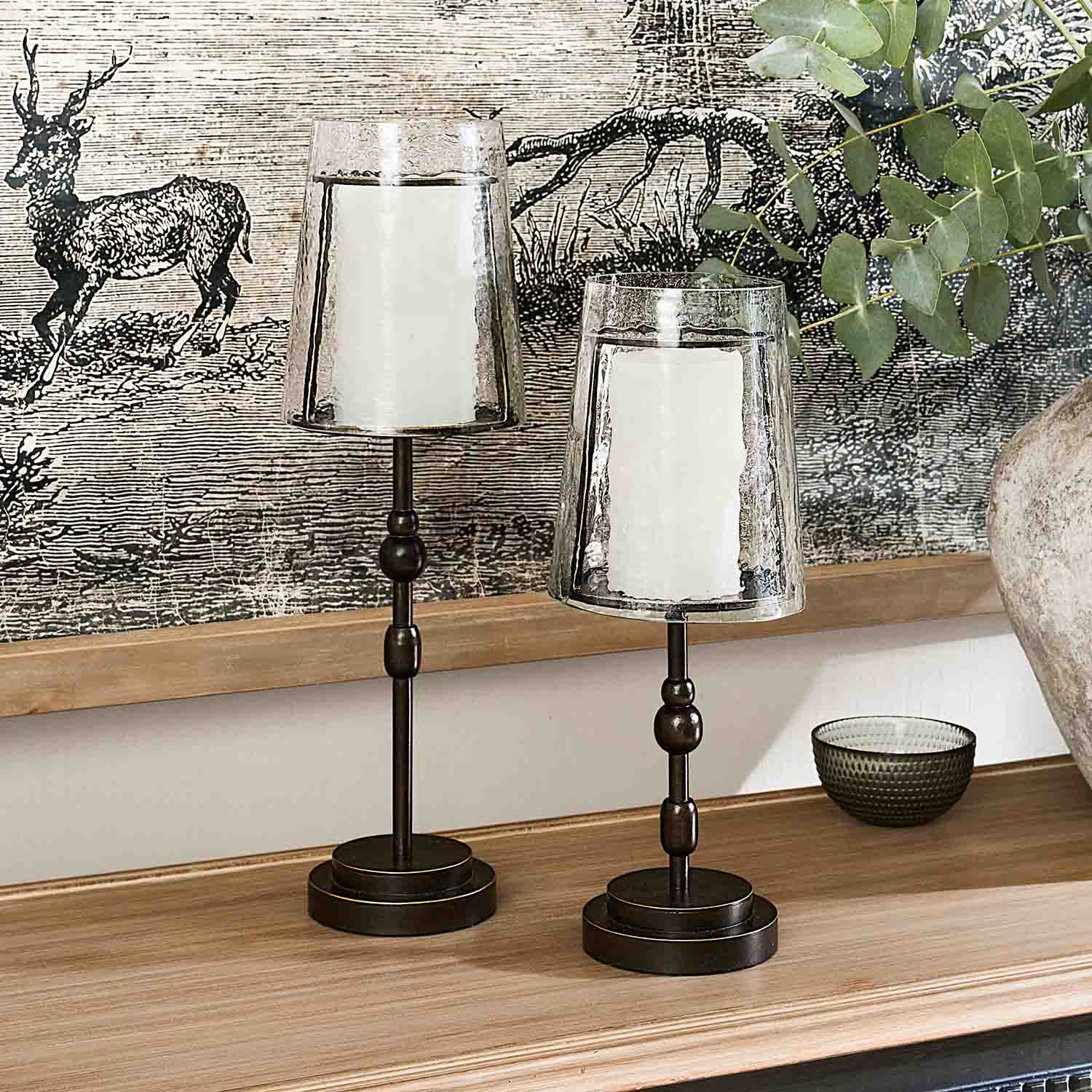 Dos candelabros de metal con pantallas de vidrio transparente y velas blancas sobre superficie de madera