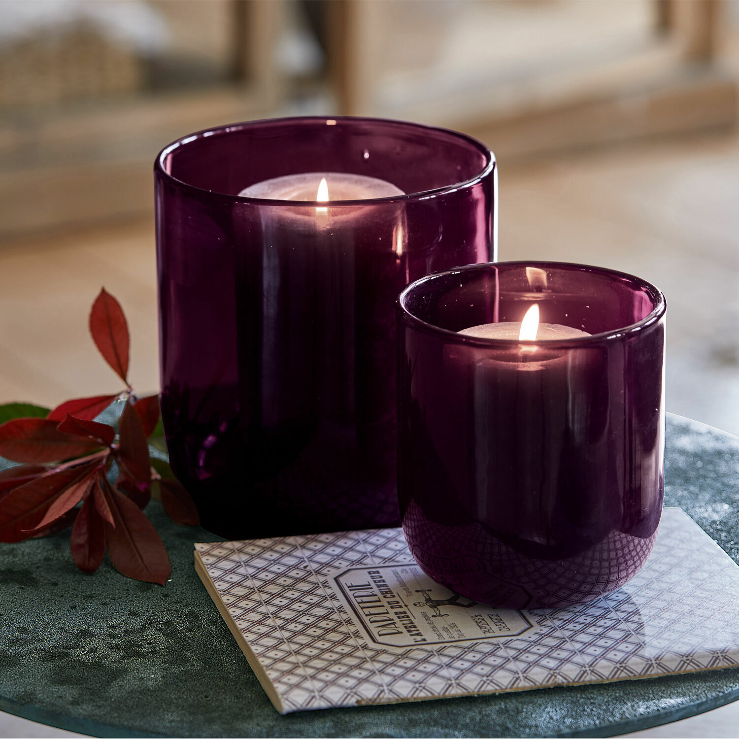 Set de 2 farolillos Theodor Dos portavelas de vidrio morado con velas encendidas sobre un soporte decorativo en mesa verde