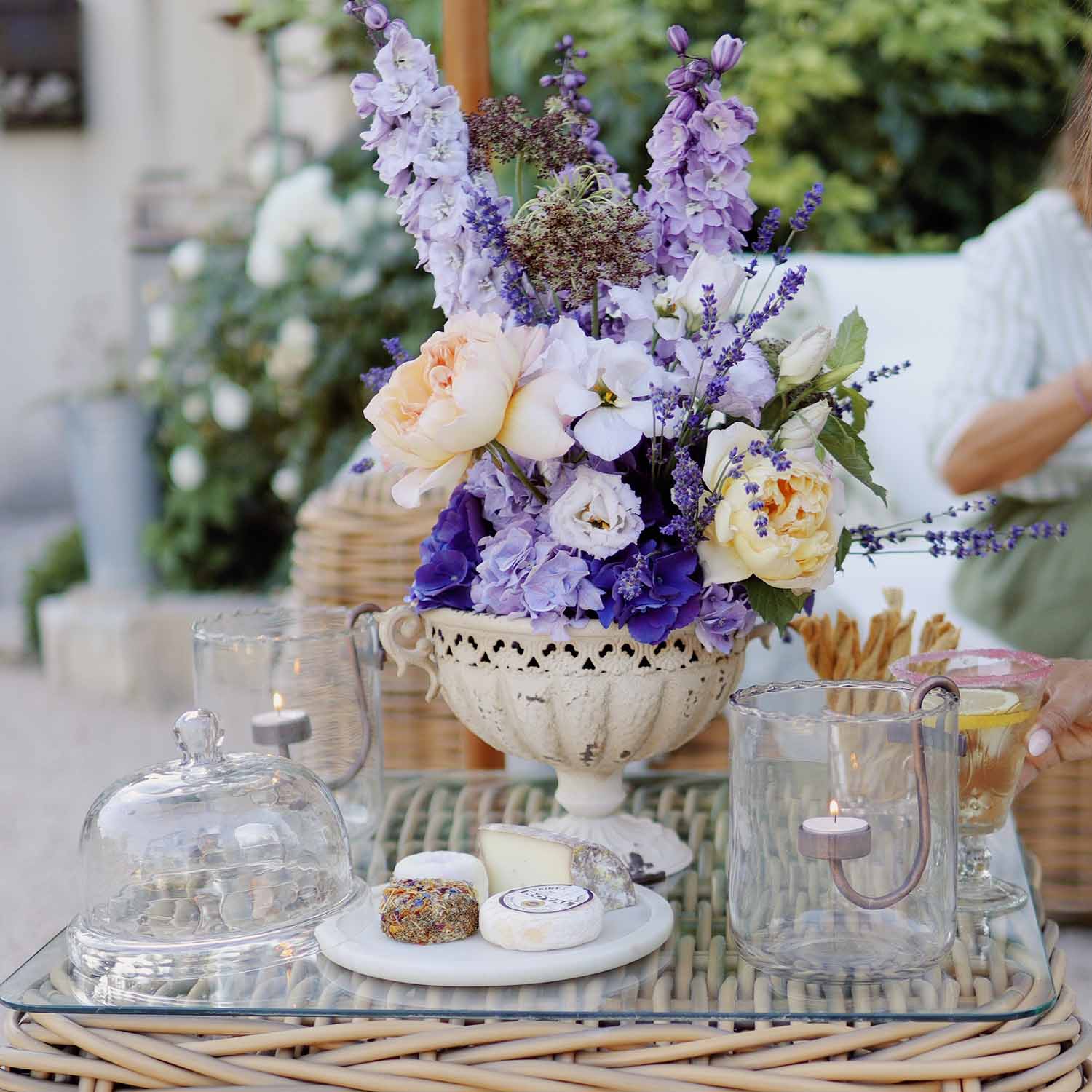 Ramo de flores violetas y crema en jarrón de cerámica blanca sobre mesa de cristal con velas y tabla de quesos