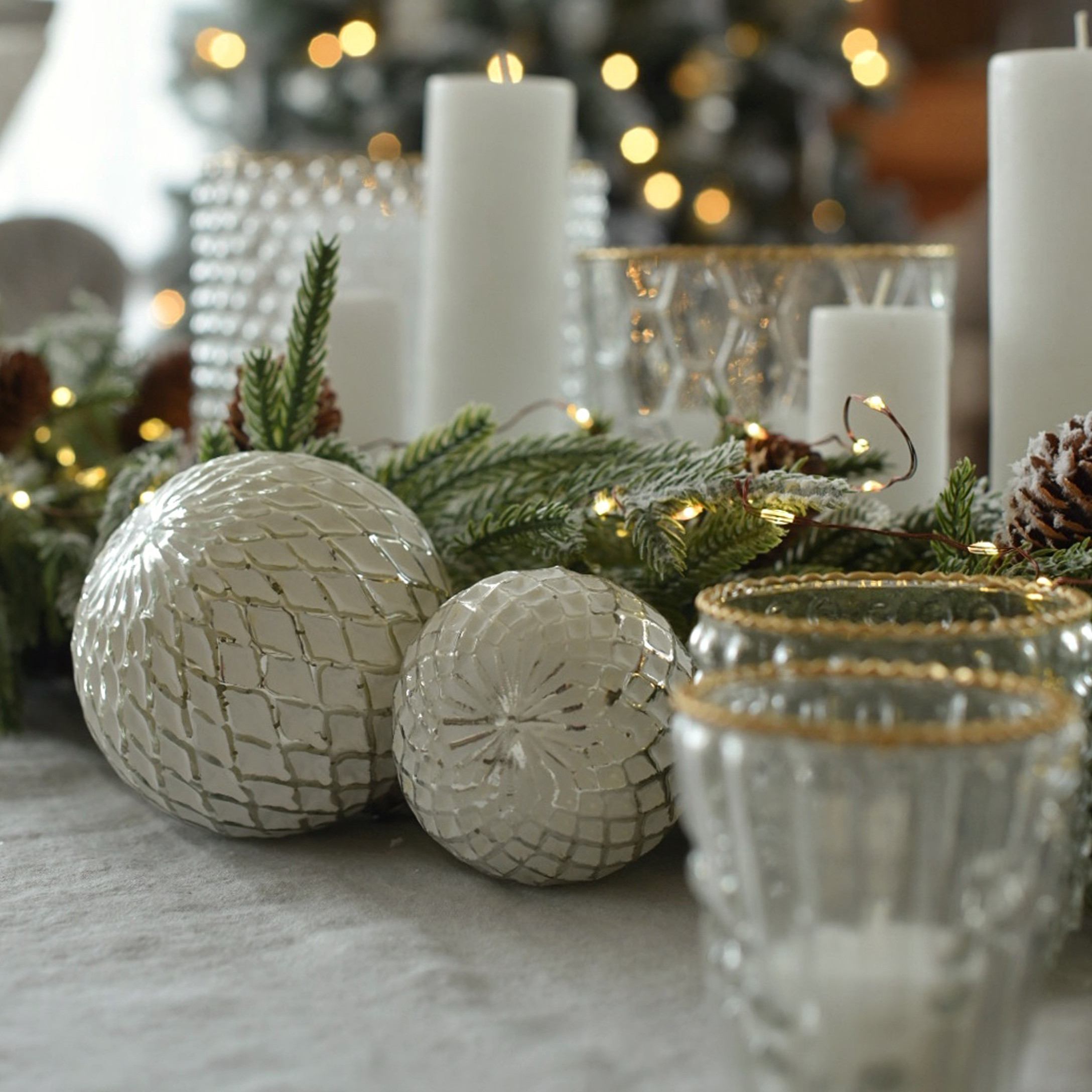 Bolas decorativas blancas de vidrio con patrón de rombos en mesa navideña con ramas verdes y velas