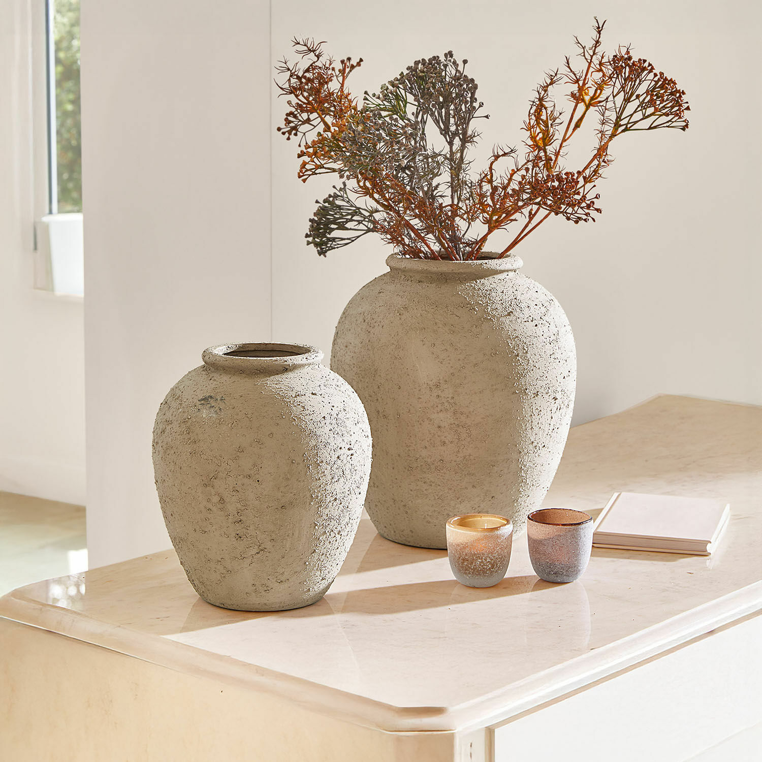 Dos grandes jarrones de hormigón gris sobre una mesa clara con ramas secas y velas decorativas