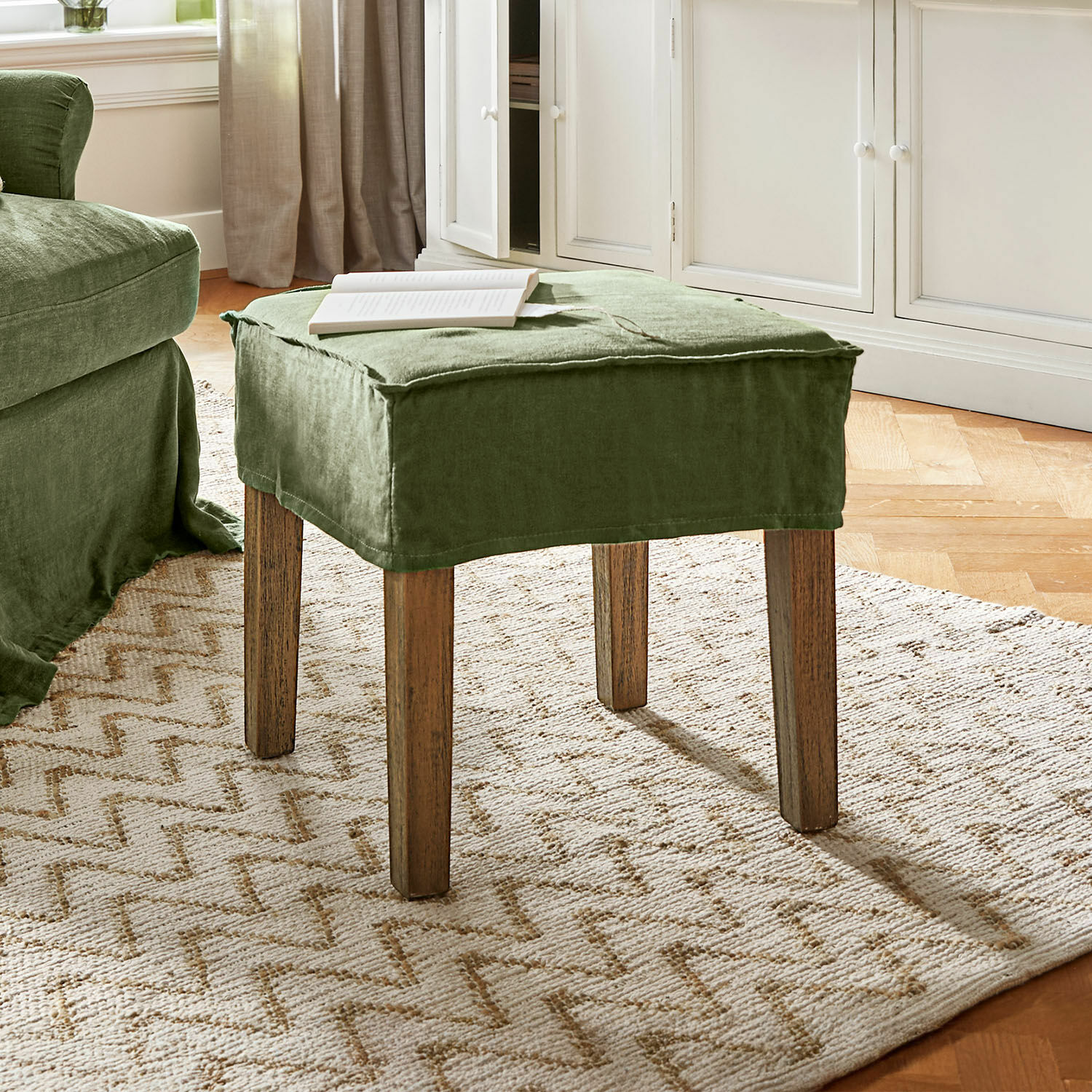 Taburete Vionnique Reposapiés verde de lino con patas de madera sobre alfombra clara en sala de estar