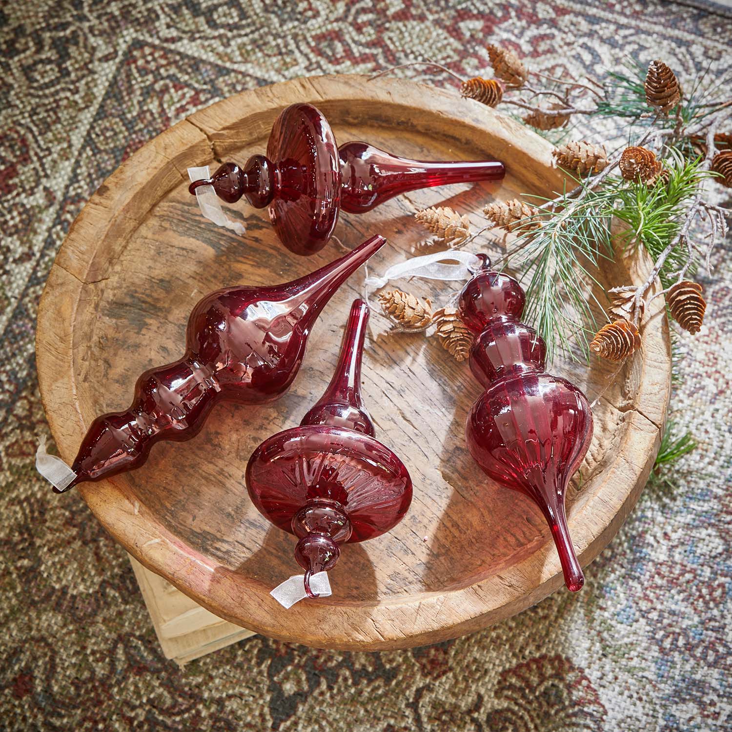 Cuatro adornos navideños de vidrio rojo sobre bandeja de madera con ramas y piñas