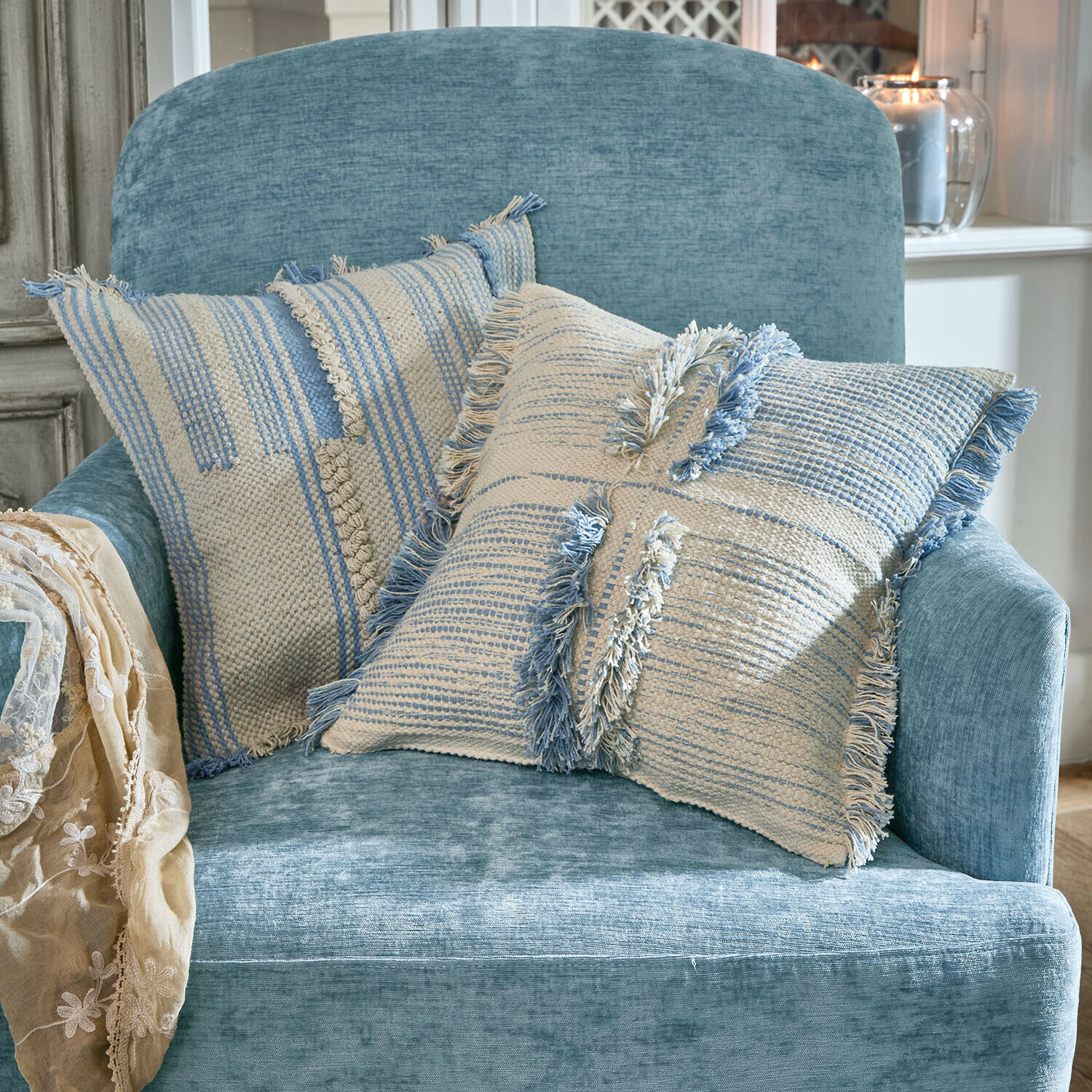Set de 2 fundas de cojín Ciel d'Azur Dos cojines decorativos de algodón en tonos crema y azul sobre un sillón azul suave en sala luminosa