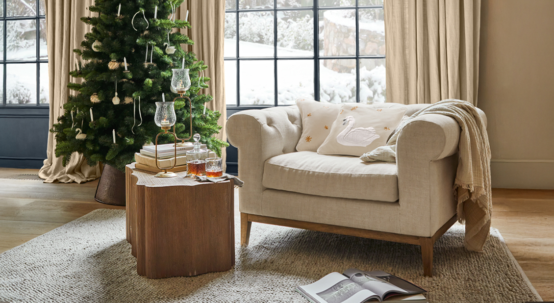 Sofá beige con cojín decorado con cisne junto a árbol de Navidad en sala acogedora de invierno