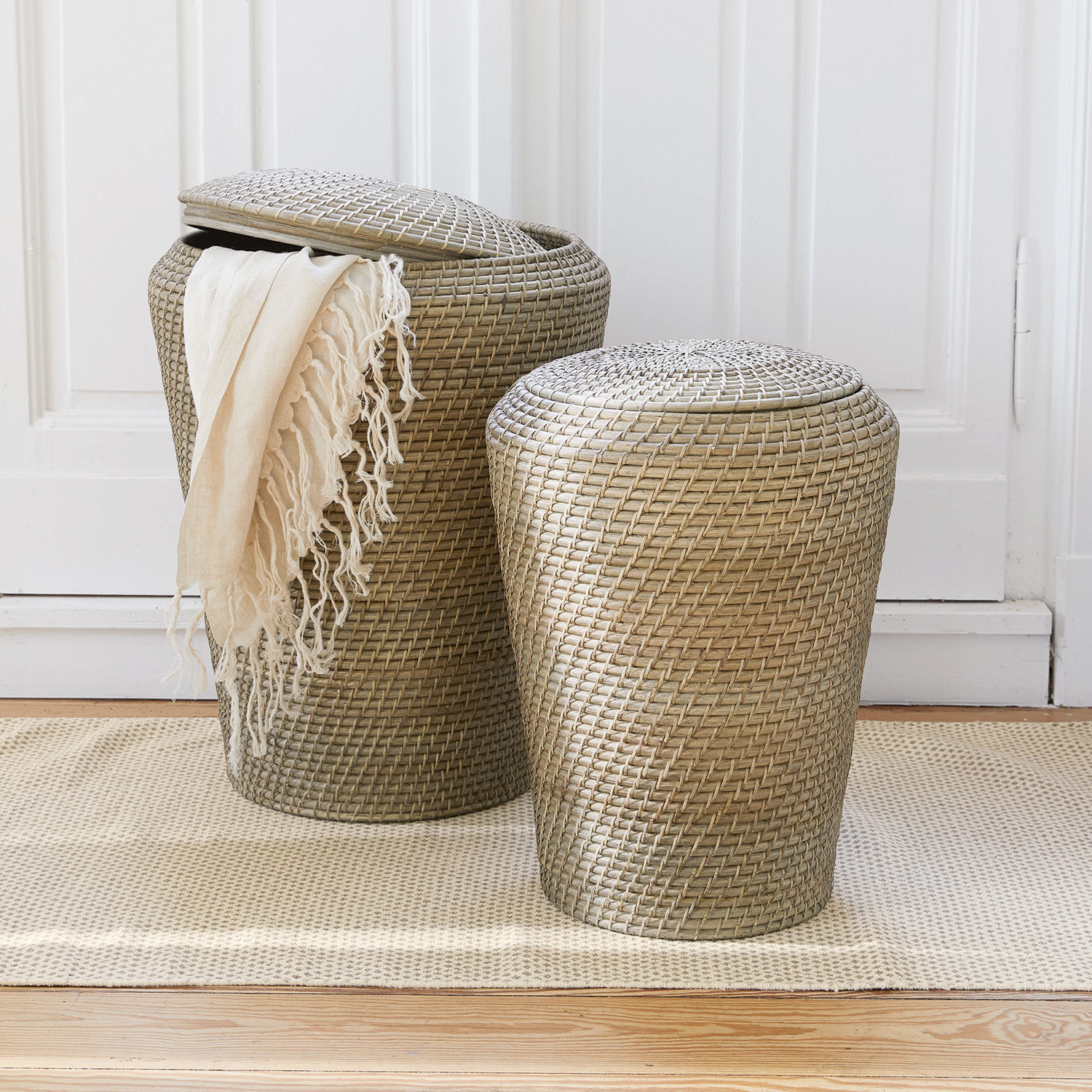 Set de 2 cestos para la ropa Nora Dos cestas de ratán beige con tapas sobre alfombra, contenedores prácticos para almacenamiento en casa
