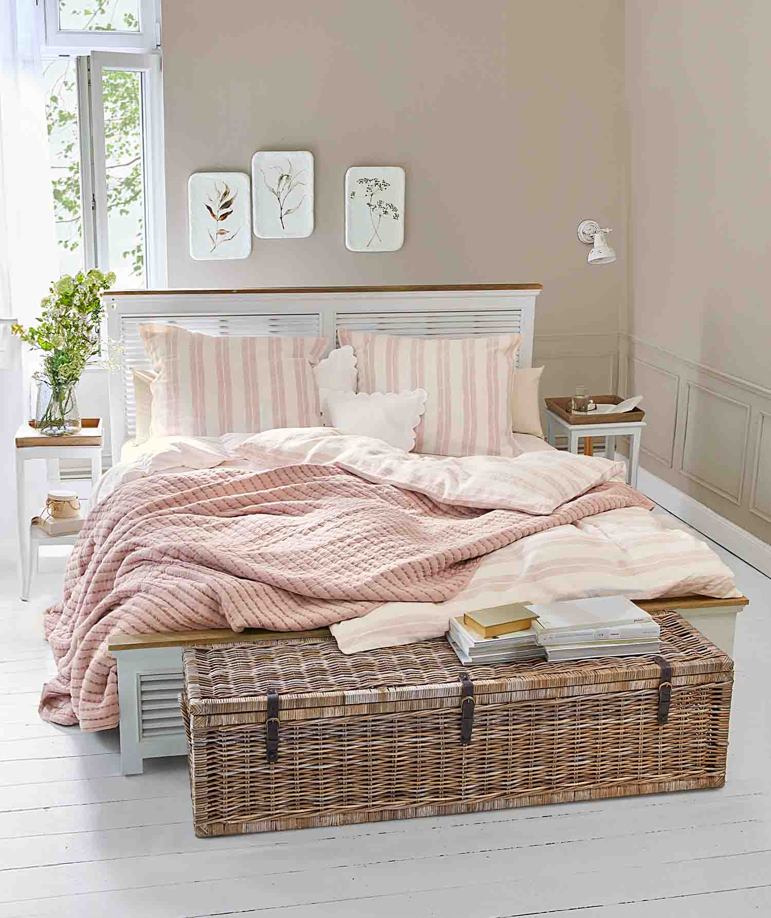 Dormitorio acogedor con cama blanca, manta rosa acolchada y baúl de mimbre con libros encima