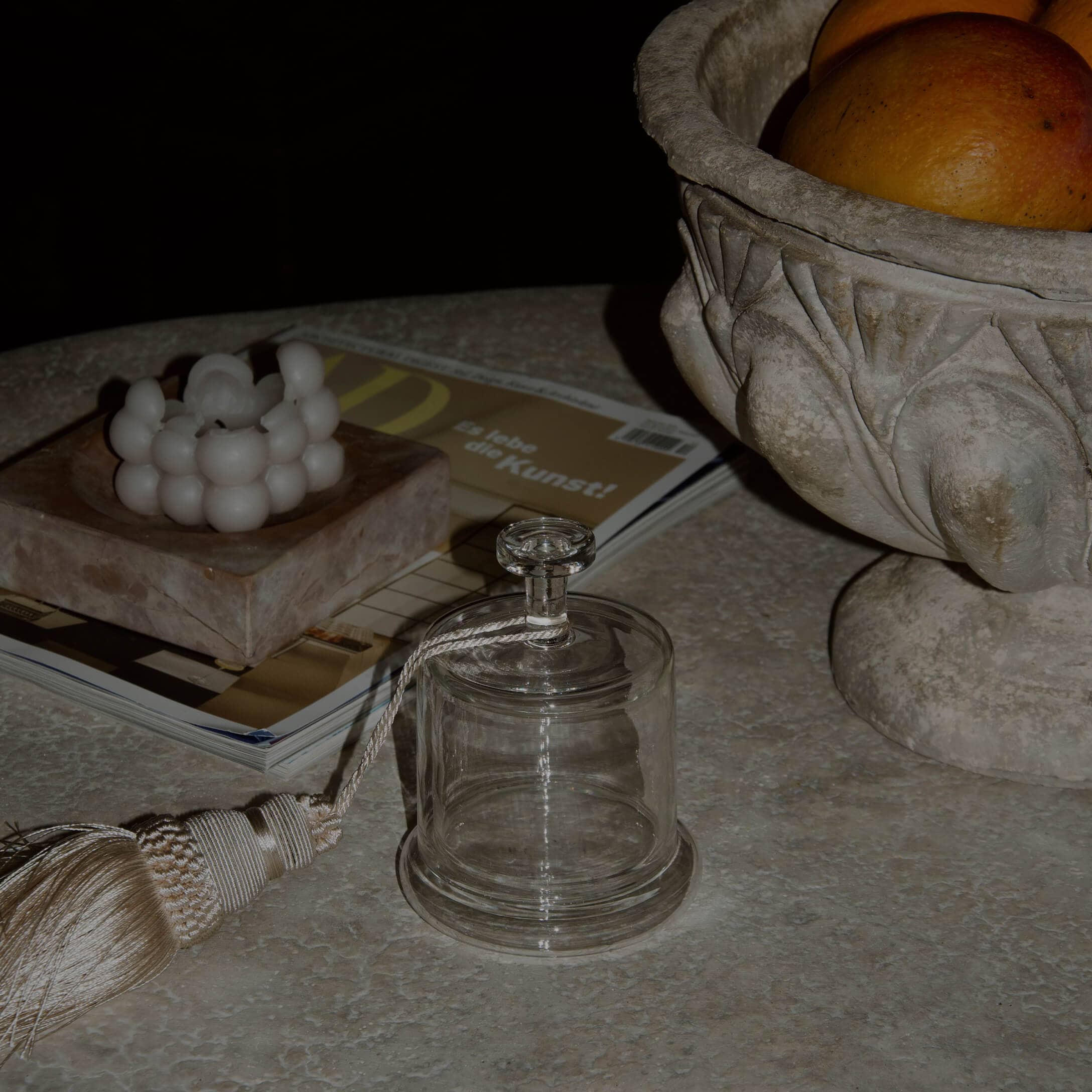 Campana de cristal transparente con borla decorativa sobre mesa de mármol con vela y cuenco de piedra