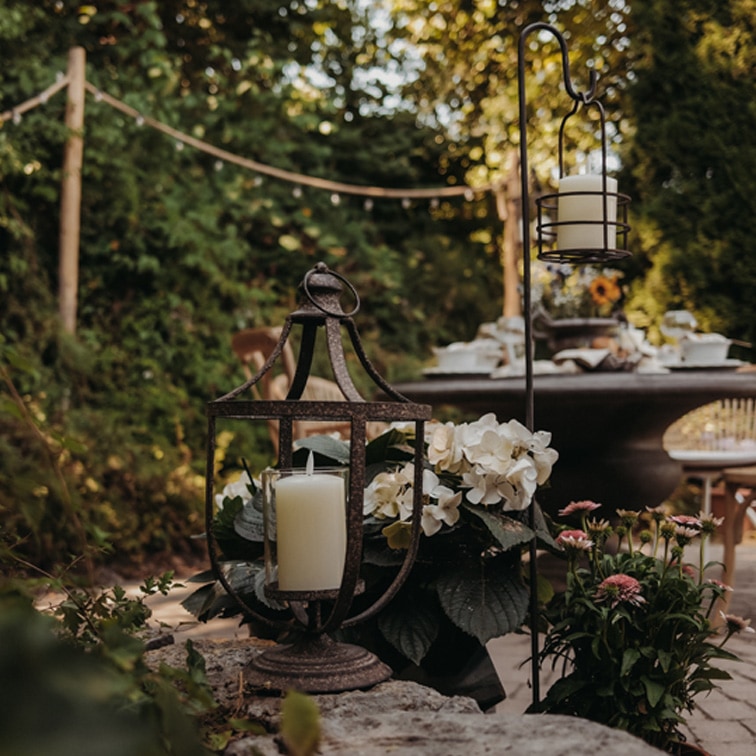 Farol de metal con vela blanca sobre muro de piedra en jardín con flores y mesa al fondo