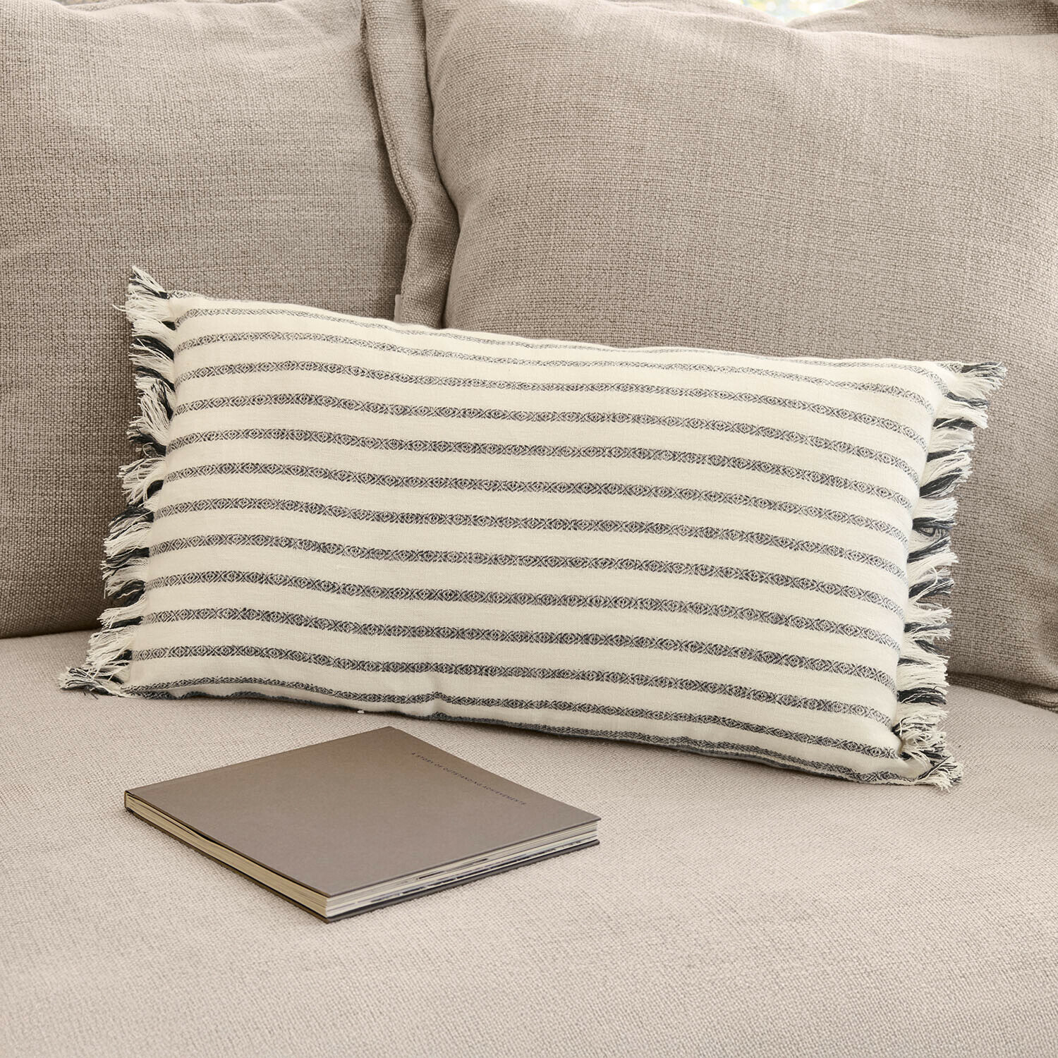 Cojín rectangular de algodón con rayas crema y gris sobre sofá beige con libro