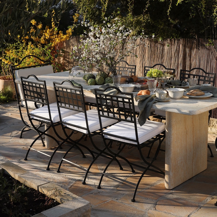 Mesa de jardín de piedra con sillas de metal negro y cojines blancos, decorada para comida al aire libre