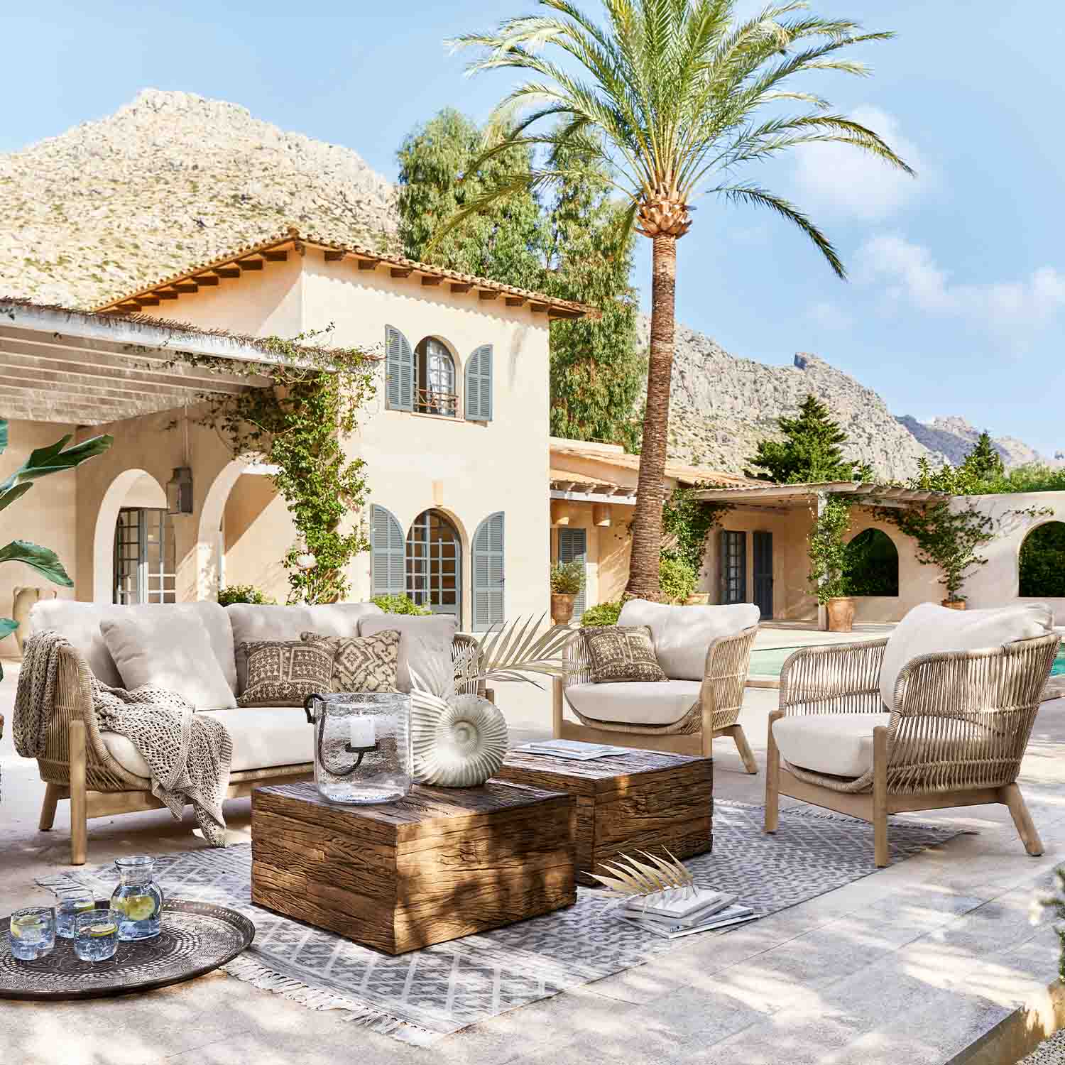 Conjunto de muebles de exterior de ratán claro con cojines beige en terraza con palmera y vistas a la montaña