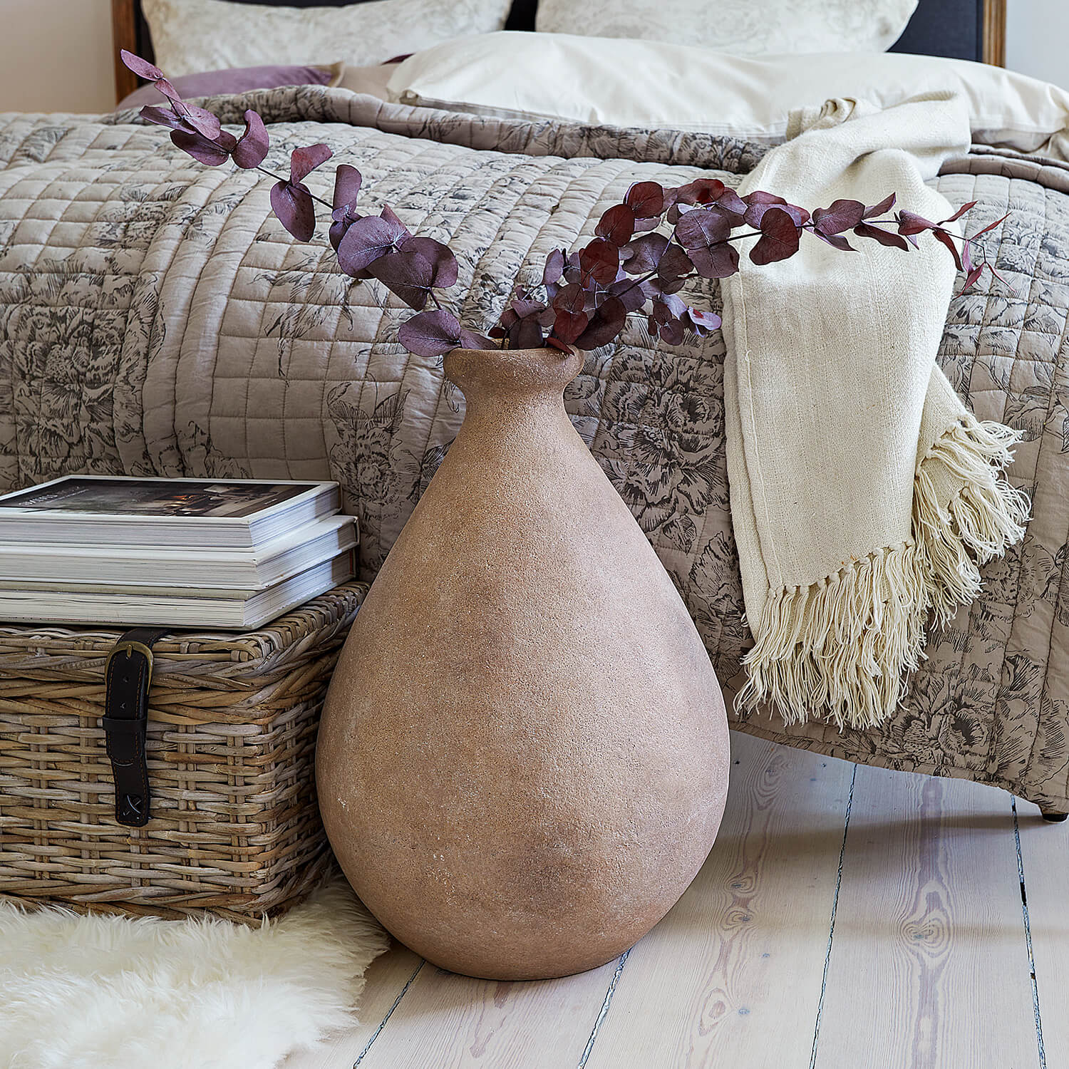Jarrón grande de cerámica beige con ramas secas frente a cama y cesta de mimbre