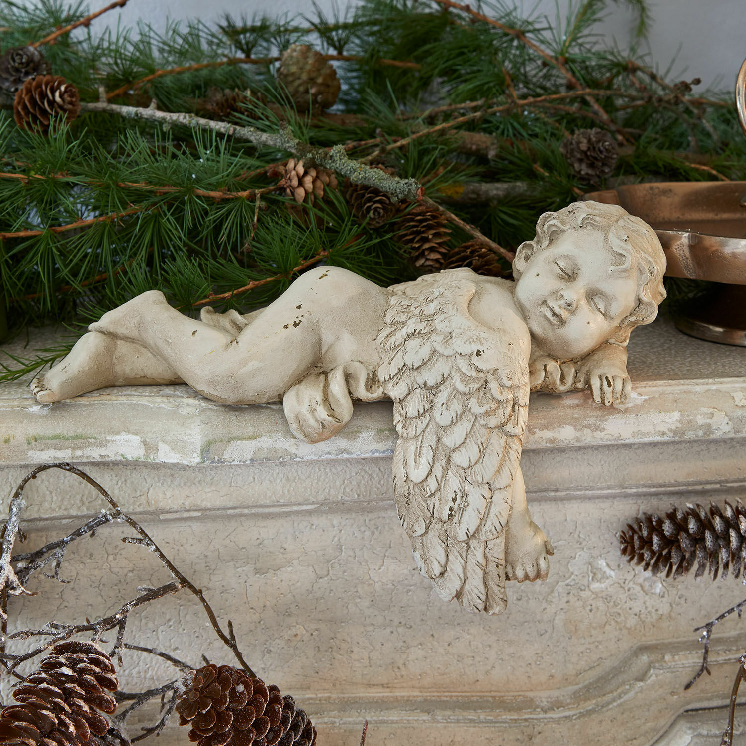 Ángel decorativo Deronza Figura de ángel de hormigón color crema sobre repisa con ramas de pino y piñas, decoración navideña