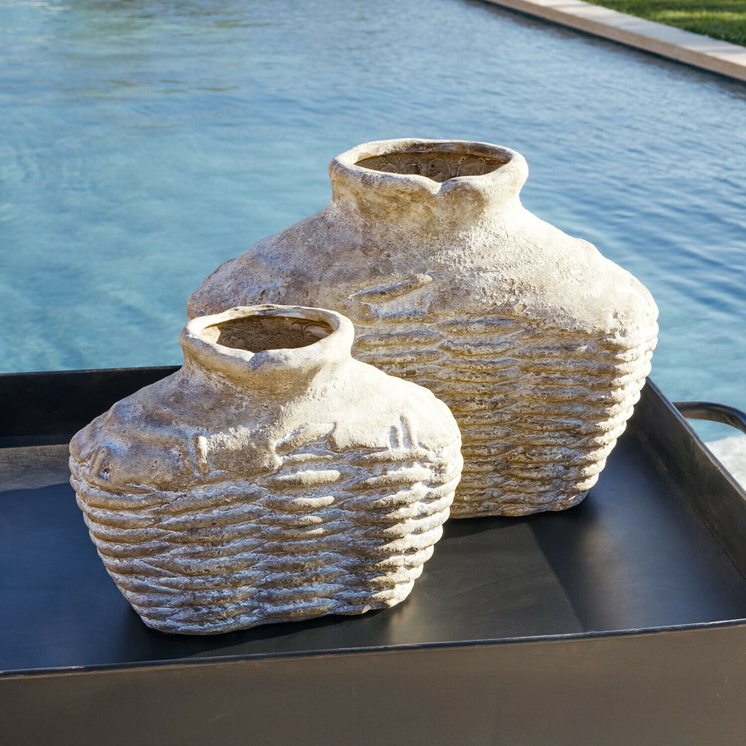 Dos jarrones decorativos de piedra beige sobre bandeja negra junto a una piscina