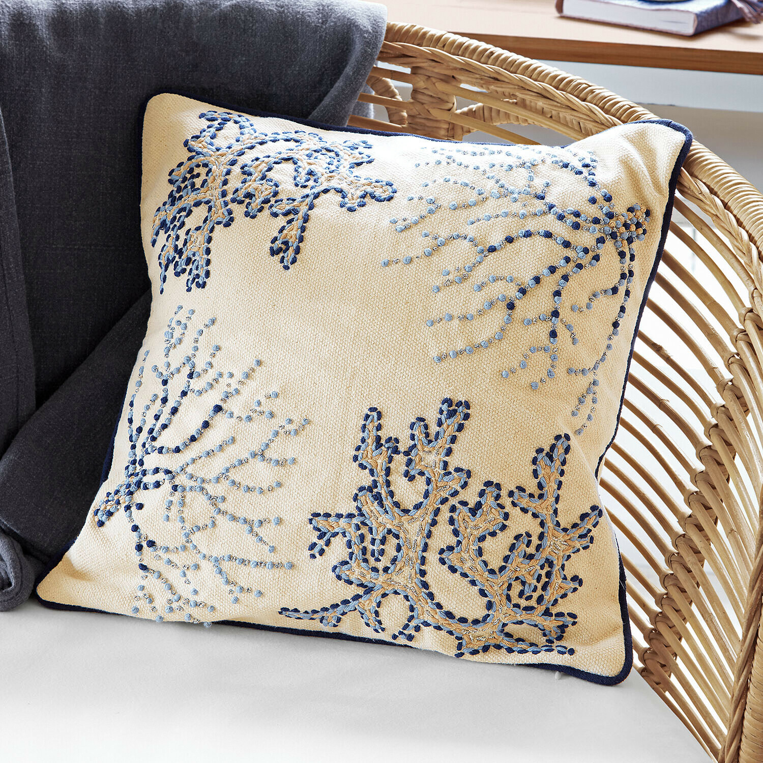 Funda de cojín Whitmere Cojín decorativo beige con bordado azul y ribete azul marino sobre sillón de mimbre