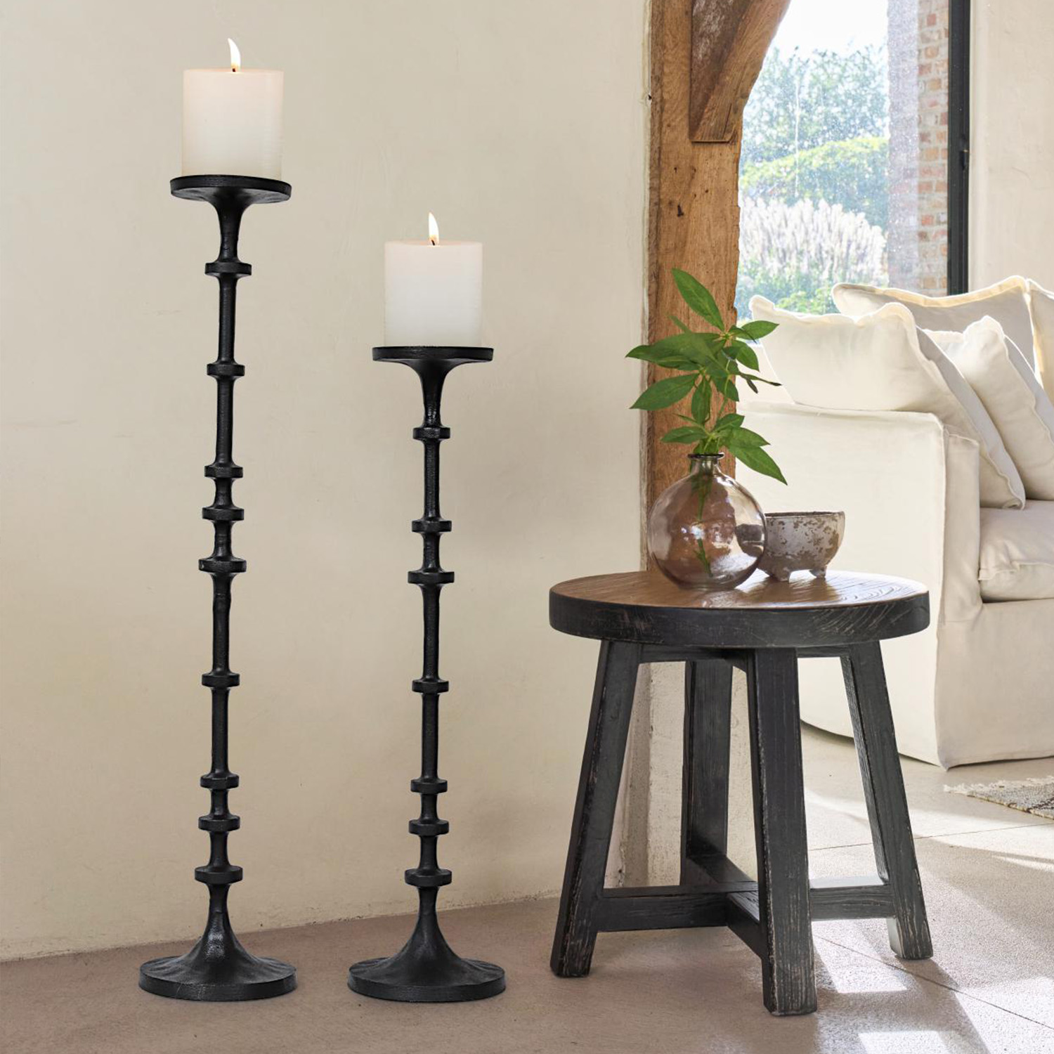 Dos candelabros de metal negro con velas blancas junto a un taburete de madera y un sofá en la sala