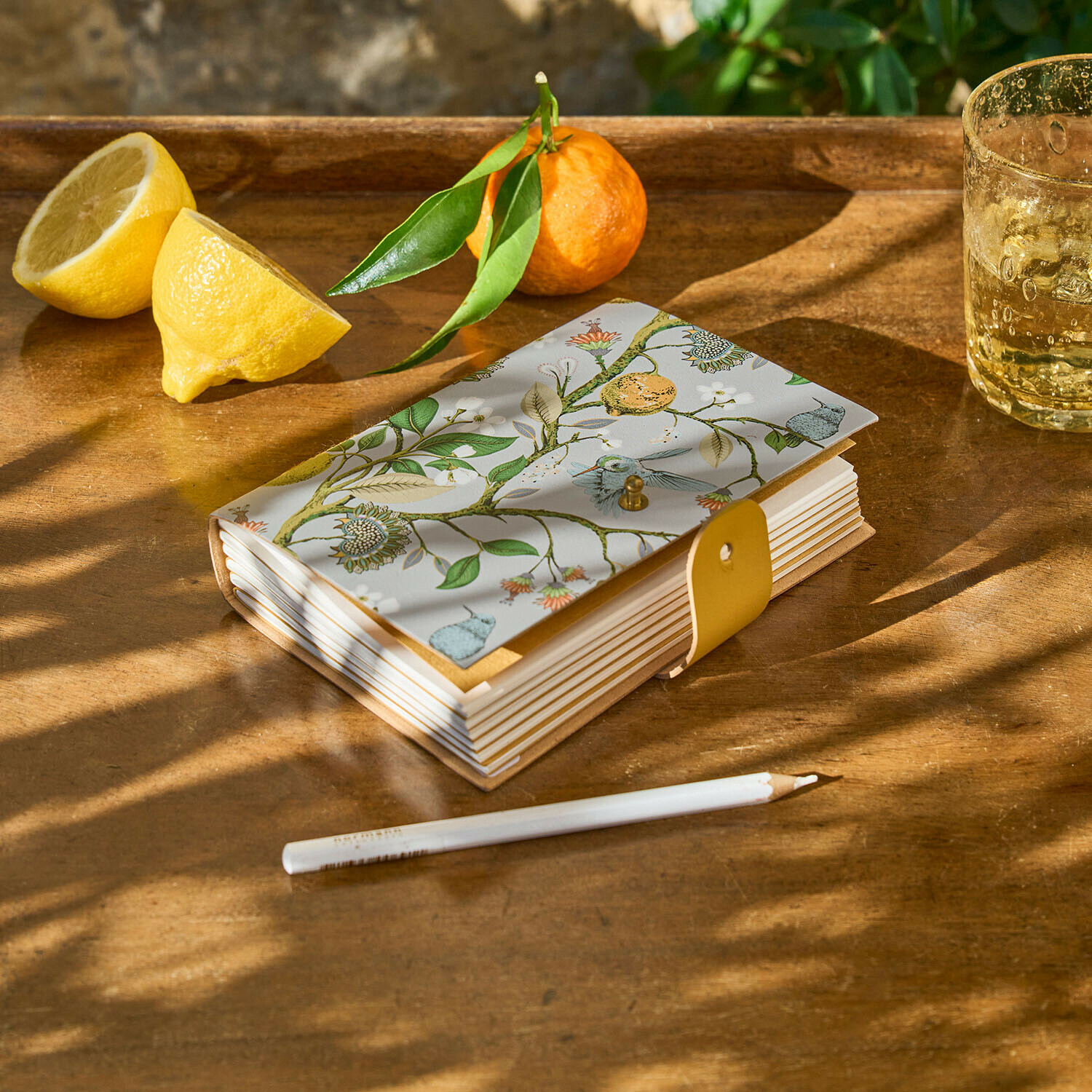 Cuaderno con cubierta decorativa de motivos vegetales y cierre amarillo sobre mesa de madera con limón y mandarina