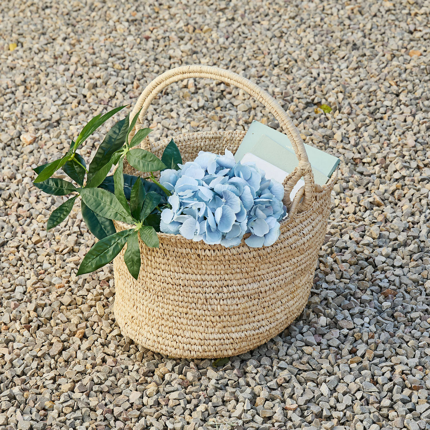 Cesta Shanoura Cesta trenzada natural de hierba marina con flores azules y hojas verdes sobre piedras pequeñas