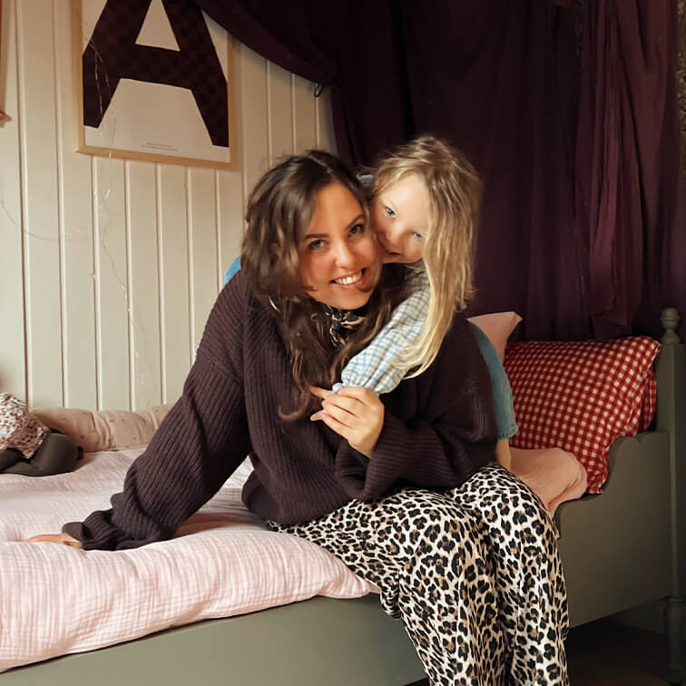 Mujer con suéter marrón y pantalones de leopardo sentada en cama con niño con camisa clara a cuadros