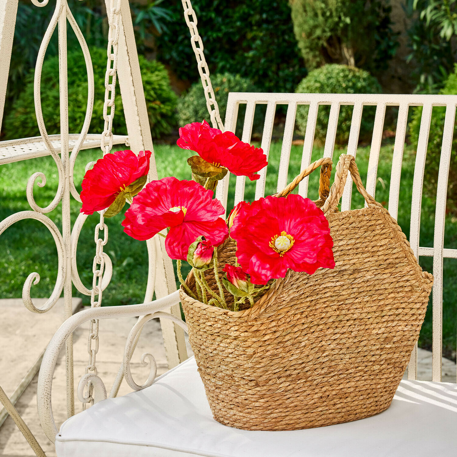 Cesta trenzada natural de hierba marina con flores rojas sobre silla blanca de jardín