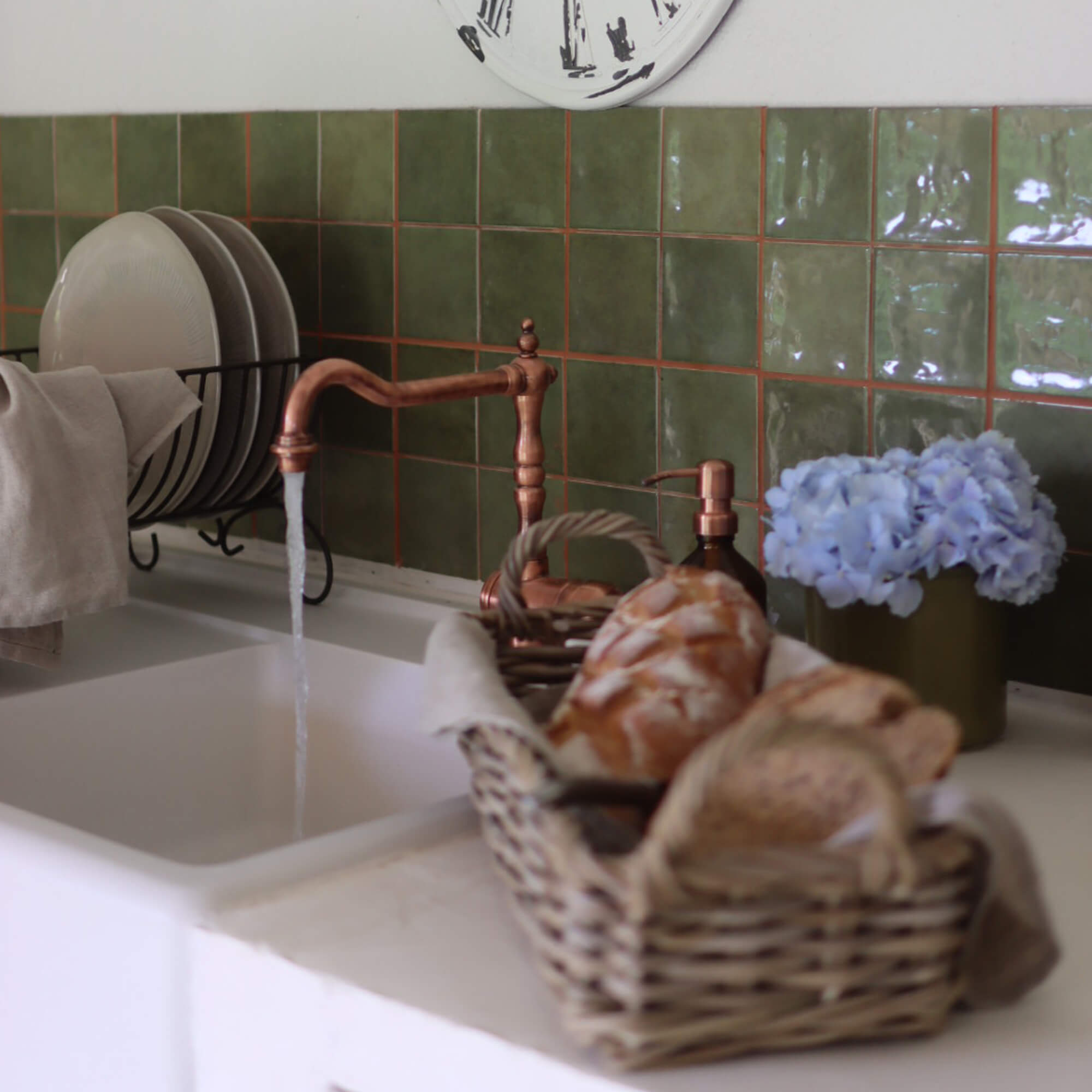 Cocina con grifo de cobre, azulejos verdes, cesta de pan y flores azules decorativas