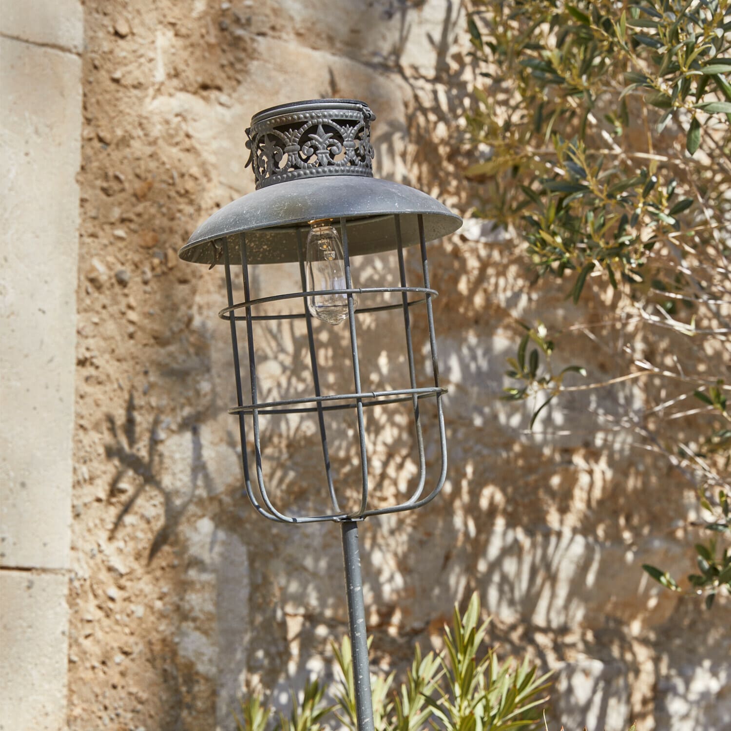 Farol solar Nistos Lámpara de jardín de metal negro con pantalla decorativa y bombilla visible sobre pared de piedra