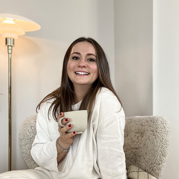 Mujer sonriente con suéter blanco sosteniendo una taza beige, sentada en un sillón suave en interior luminoso
