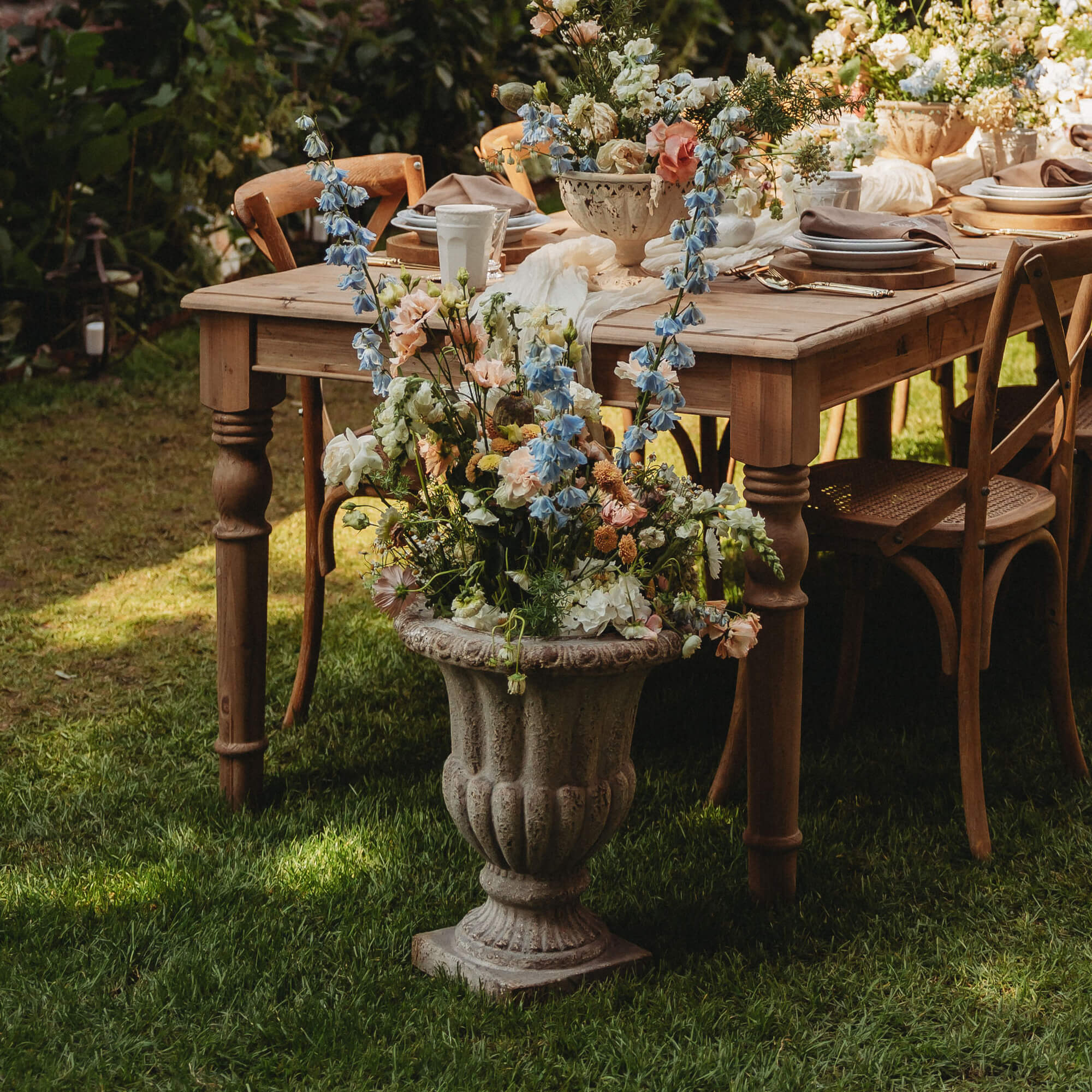Gran jarrón de piedra con flores en tonos pastel frente a mesa y sillas de madera en jardín