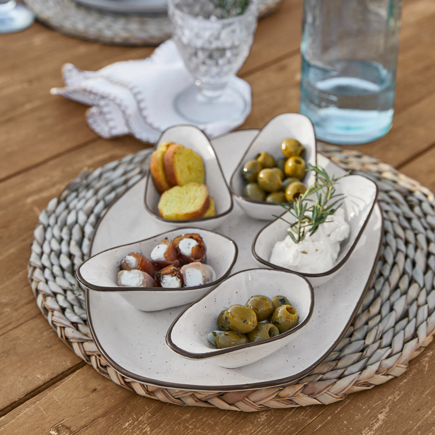 Set de 5 cuencos y plato de servir Biarré Pequeños cuencos de cerámica blanca con aceitunas y queso sobre base trenzada de madera